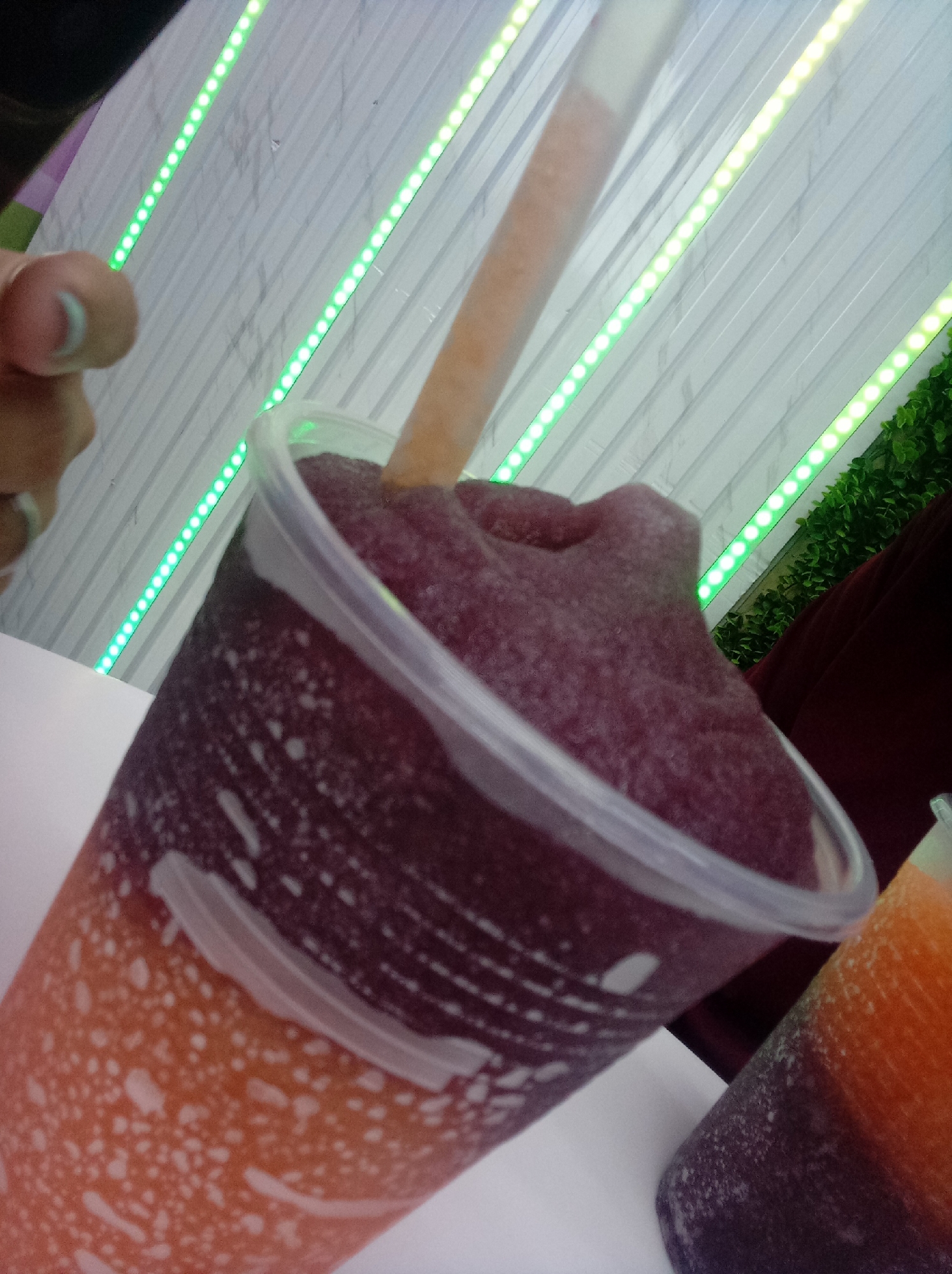 The image shows two layered frozen drinks in clear plastic cups with straws, one prominently in the foreground and a second partially visible to the lower right. The drink in the foreground has a layered effect, transitioning from orange at the bottom to dark purple at the top. There are visible condensation and streaks on the exterior of the cups. A hand with painted fingernails is partially visible on the left side of the frame. The background consists of a white, textured wall with linear green LED lights running diagonally across it. Some artificial greenery is also visible in the upper-right background. The scene appears to be indoors, possibly a food stall or shop. There is no evidence to confirm the location is in the city of Guarenas, Venezuela.