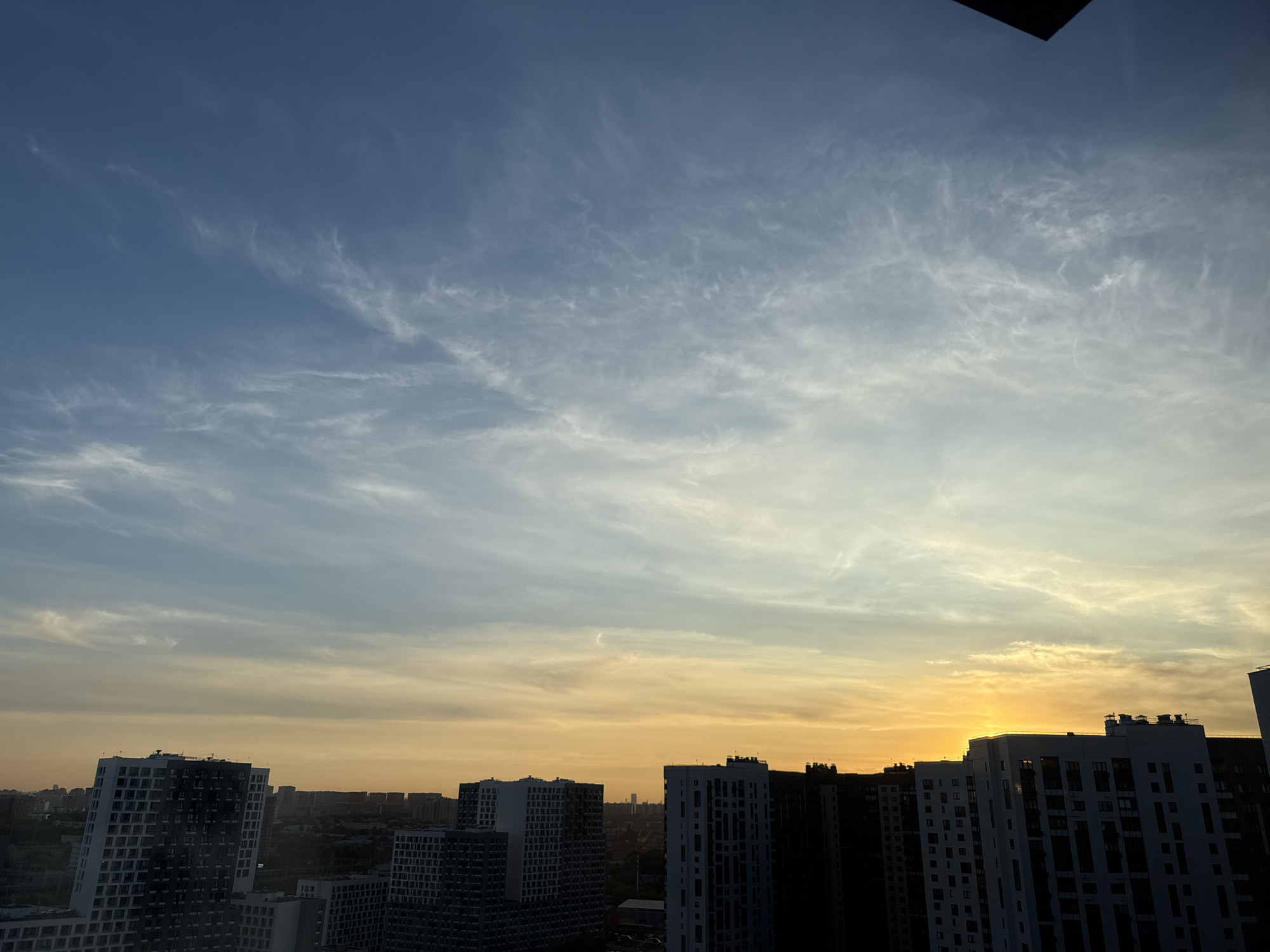 The image captures a sunset over the city of Lyubertsy, Russia. A gradient of blue and yellow fills the sky, transitioning from a clear blue at the top to a bright yellow near the horizon, where the sun is setting behind a line of apartment buildings. Wispy white clouds are scattered across the sky, particularly in the upper portion. The foreground is dominated by the silhouetted shapes of modern, multi-story apartment buildings, some with distinctive architectural features like angled facades and numerous windows. The buildings are dark, indicating that the exposure is focused on the brightly lit sky. The overall impression is of an urban landscape at dusk. There are no people or identifiable actions depicted in the image.