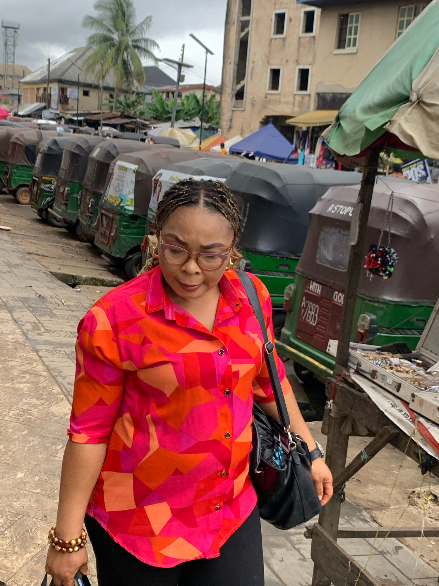 In Umuode, Nigeria, a woman with braided hair and glasses walks along a paved path next to a row of parked auto-rickshaws. She wears a bright, multi-colored geometric print blouse and black pants, carrying a black handbag on her shoulder. A beaded bracelet adorns her left wrist. Behind the auto-rickshaws, market stalls are visible, with goods displayed on tables and covered by umbrellas. Buildings and a palm tree are in the background, under an overcast sky.