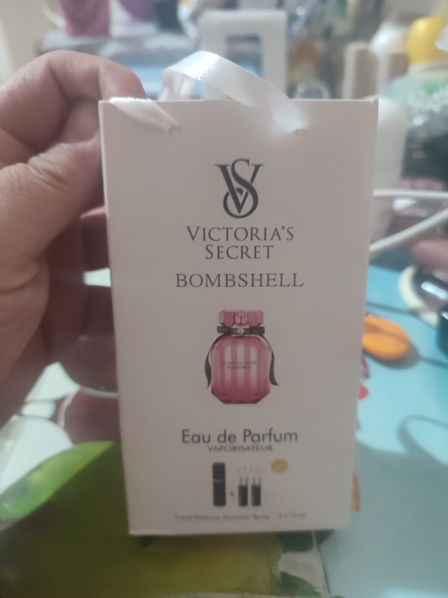 A hand holds a white cardboard box with a ribbon handle. The box has a Victoria's Secret logo at the top, followed by the words "VICTORIA'S SECRET" and "BOMBSHELL" in black text. Below this is an image of a pink striped perfume bottle. At the bottom of the box, the text "Eau de Parfum VAPORISATEUR" is visible, along with images of three small perfume bottles. The background is blurred, showing what appears to be a shelf with various items. The image does not contain any information that could be used to determine its location in Chernivtsi, Ukraine.