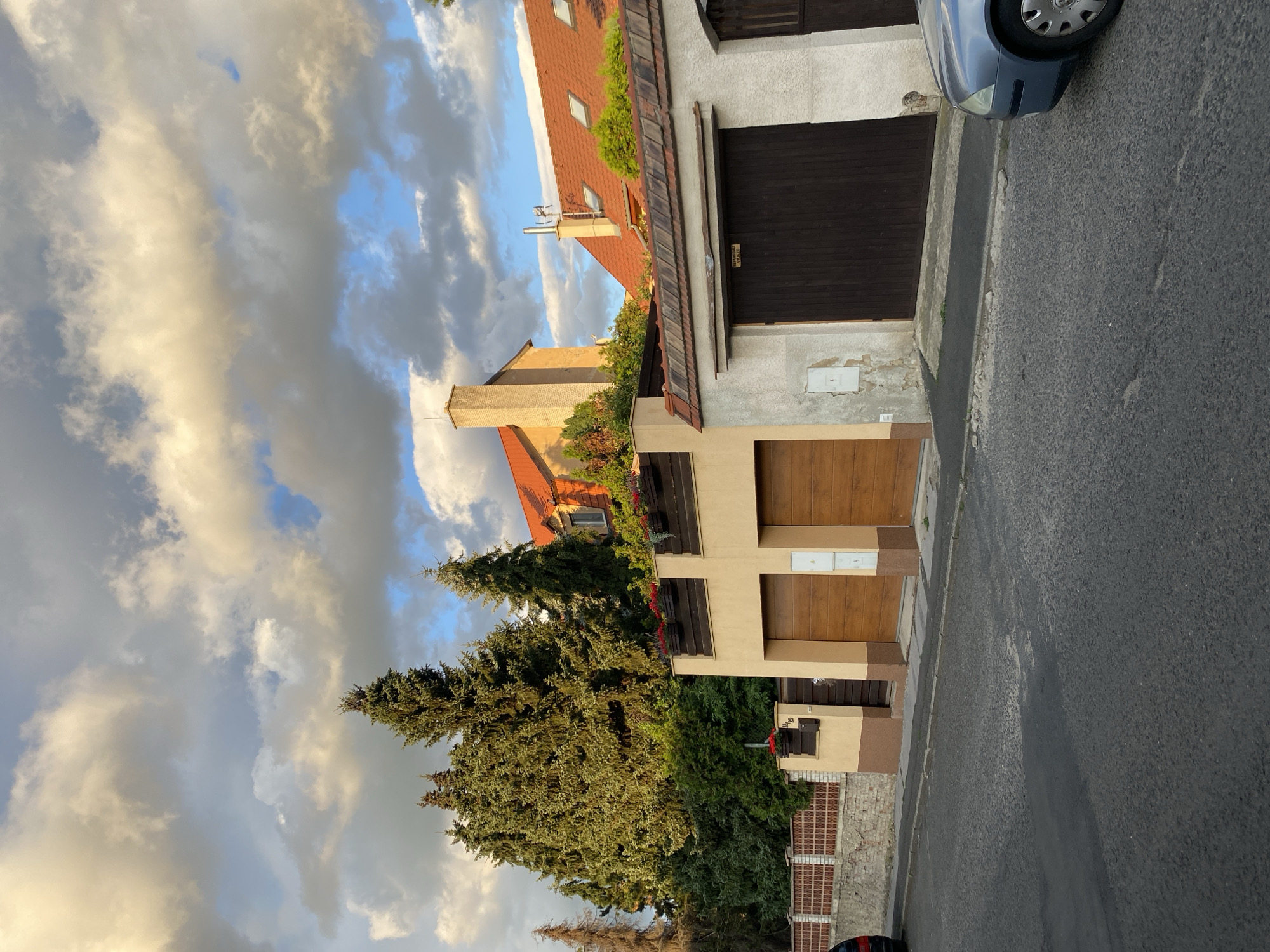 The image shows a residential street in Bílina, Czechia. On the right side of the street is a row of houses with red tile roofs. The houses have garages with wooden doors. The street is paved with asphalt. A blue car is parked on the right side of the street. In the background, there are tall trees. The sky is cloudy with patches of blue sky visible. The scene appears to be captured during late afternoon or early evening, as indicated by the warm lighting and the shadows.
