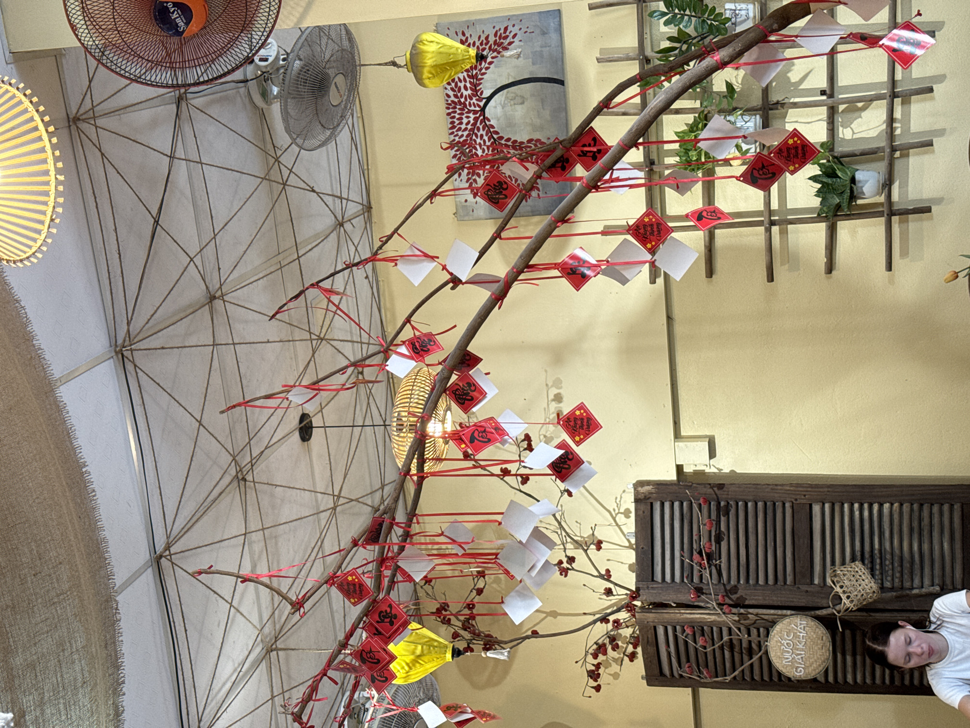 The image captures a festive interior scene, likely a cafe or restaurant in Hoi An, Vietnam, decorated for a holiday, possibly Tet. The ceiling is adorned with a woven bamboo lamp and a ceiling fan. A prominent branch, decorated with red ribbons and small red diamond-shaped ornaments with black calligraphy, is suspended from the ceiling. White square papers are also hung from the branch. In the background, a yellow lantern hangs, and a painting of a red tree is visible. To the left, a wooden shuttered cabinet stands, with a woven sign reading "NƯỚC GIẢI KHÁT" (Beverage) and a small woven basket. A young woman with blonde hair, wearing a white t-shirt, is seated on the left, looking down. The overall color palette is warm, with yellow walls and brown wooden elements contrasting with the red and white decorations.