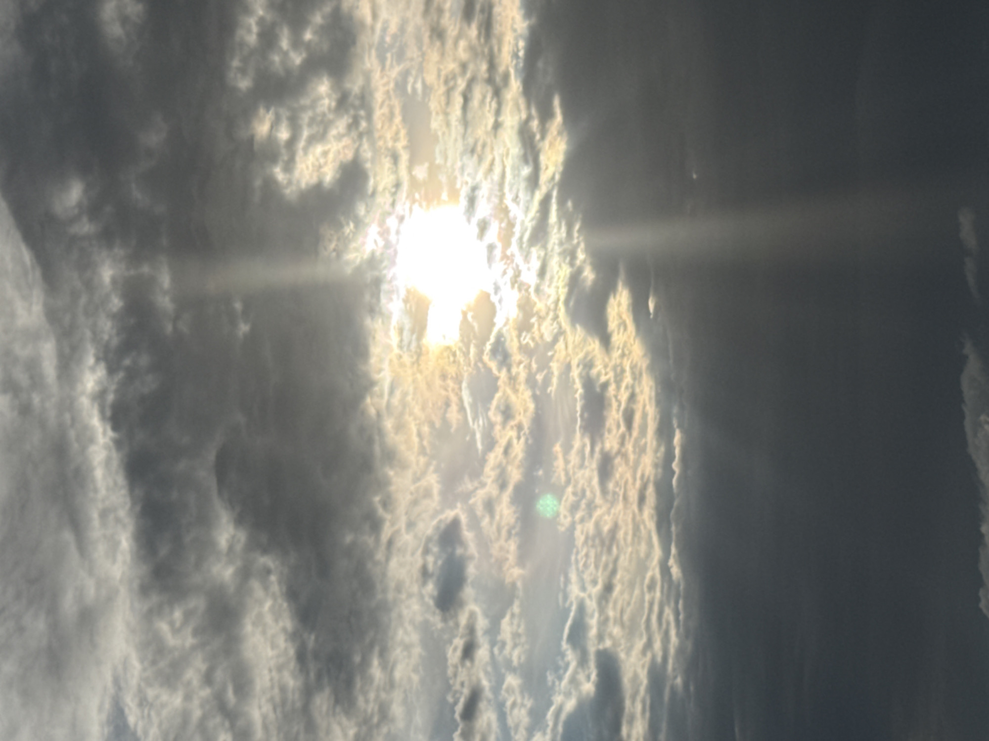 A vertical, eye-level, full shot captures a dramatic sky filled with sunbeams piercing through clouds. The sun, a bright, white orb, is positioned slightly right of center, creating a radiant halo effect. Rays of light extend downward and to the right from the sun, illuminating the surrounding clouds. Some clouds are dark and heavy, indicating potential rain, while others are lighter and wispy, allowing the sunlight to shine through. There are no people, animals, or man-made objects visible in this natural landscape. The overall impression is one of a powerful and dynamic sky, likely taken during sunrise or sunset due to the dramatic lighting.