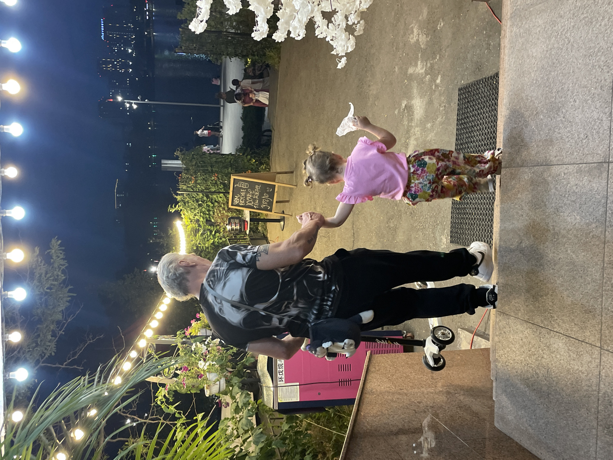 A man and a young girl are walking down a stone staircase at night. The man, who is on the left and slightly ahead, wears a black t-shirt with a skull graphic and black pants. He is holding the girl's hand. He has a small stuffed animal hanging from his back pocket. The girl, on the right, wears a pink t-shirt and colorful patterned pants. She is holding a white object in her right hand. They are on a paved area next to some greenery and outdoor lighting. In the background, there is a body of water with buildings and lights visible on the opposite shore, suggesting an urban waterfront setting, possibly in Almaty, Kazakhstan given the context.