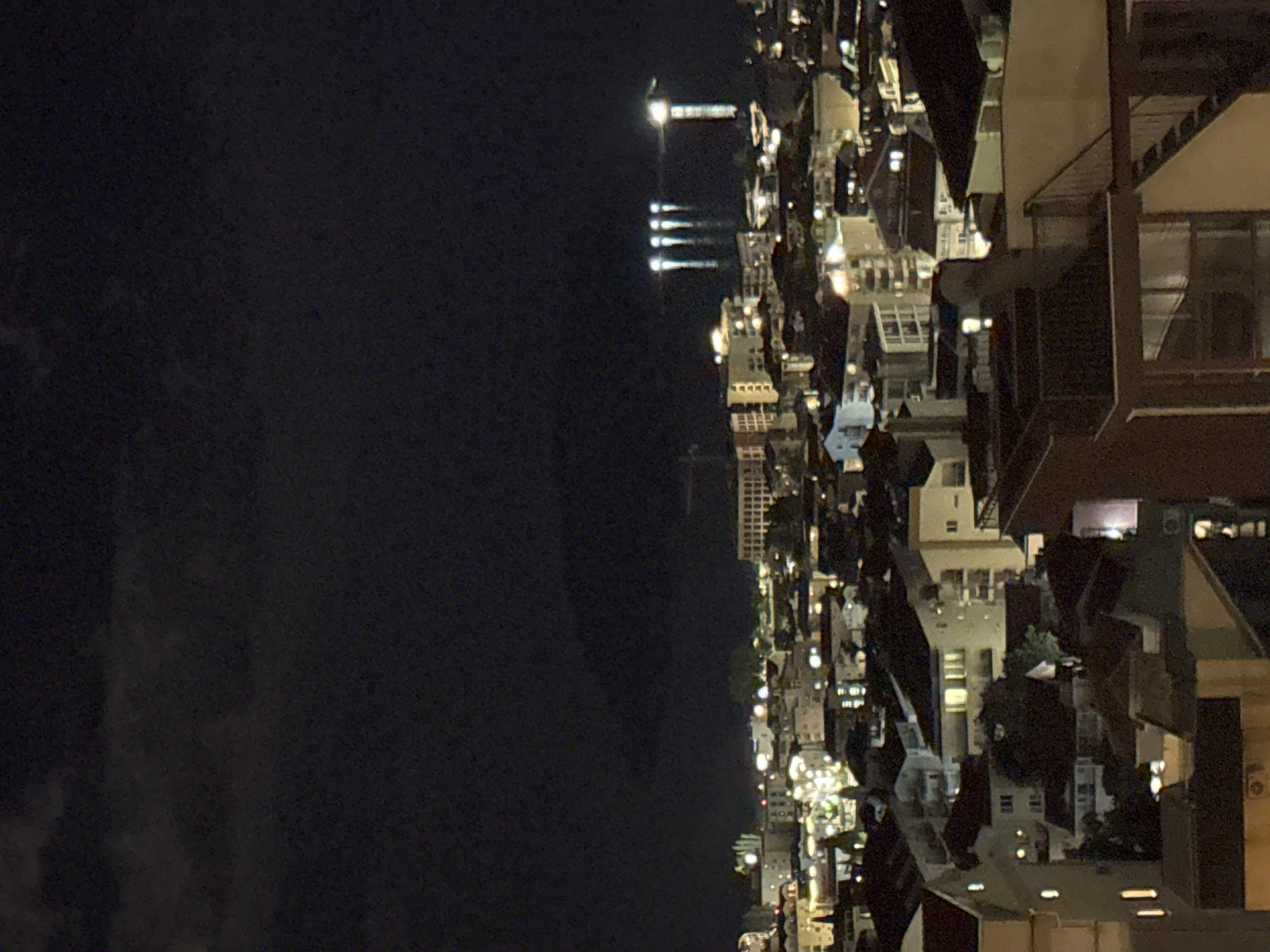 The image displays a nighttime panoramic view of a coastal city, identified as Budva, Montenegro, captured from an elevated vantage point. The scene is dominated by the artificial lights of the urban environment set against a dark sky and sea.
In the foreground and midground, a dense collection of buildings spans the landscape, illuminated by numerous windows and streetlights. These structures, including apparent residential or commercial buildings, feature balconies and varied architectural details, with many lights casting a warm, yellowish glow. Some darker patches within the city suggest unlit areas or natural terrain.
The background transitions into a vast, dark expanse, which appears to be the sea blending into the night sky. In the far distance, a prominent, bright light source is visible, resembling a lighthouse or a significant illuminated structure, possibly at the end of a pier or breakwater. Below this main light, four distinct vertical streaks of light are reflected on the water's surface, indicating the presence of a calm body of water. Faint, dark cloud formations are discernible in the upper regions of the dark background.
The time of day is clearly night, and the weather appears clear. No specific human activity is observable, nor is any discernible text present in the image. The overall impression is one of a serene, illuminated cityscape by the coast.