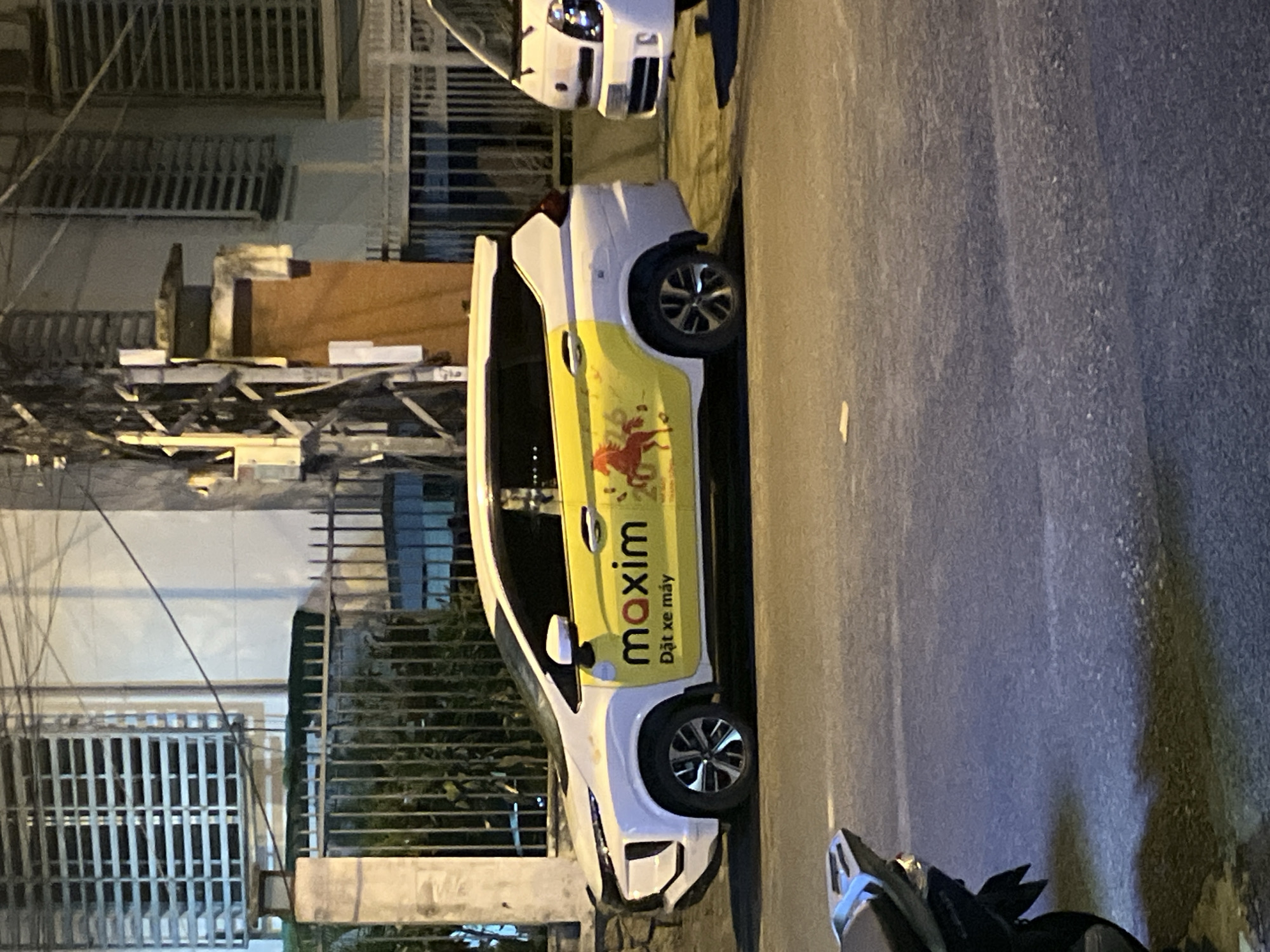 An outdoor, eye-level, medium shot shows a white Maxim ride-sharing car parked on a street at night in Nha Trang, Vietnam. The car is white with yellow branding on its side that says "maxim" in red letters, "Đặt xe máy" in smaller black letters below, and a red horse graphic. The car has tinted windows and alloy wheels. To the right of the car, a white van is partially visible. The background consists of a building with barred windows and a metal fence. The street is paved and appears wet, possibly from rain. A motorcycle is partially visible in the lower left corner of the frame. The lighting suggests it is evening or nighttime, with artificial lights casting shadows.