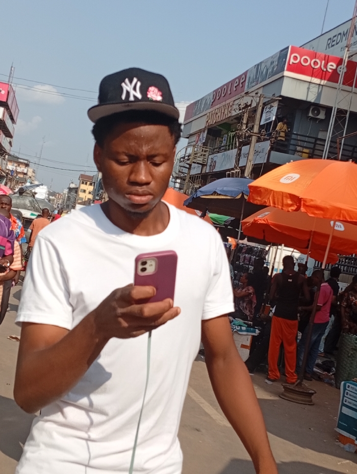 A young man wearing a white t-shirt and a black "NY" baseball cap looks at his purple smartphone, which is connected to a white wired earphone. He stands outdoors in a bustling marketplace with other people milling around. Orange umbrellas provide shade to vendors. Buildings with storefronts are visible in the background, one bearing the name "Poolee".