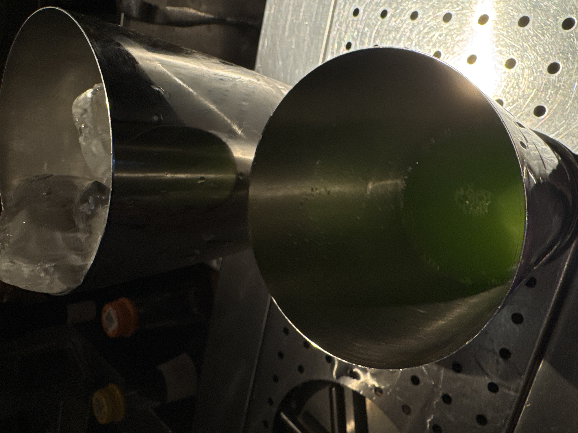 In Yerevan, Armenia, a close-up shot captures a bar scene with two stainless steel cocktail shakers. The larger shaker in the background is filled with ice and positioned slightly behind and to the right of the foreground shaker. The foreground shaker is closer to the viewer and contains a vibrant green liquid with some bubbles, suggesting a freshly prepared cocktail. The shakers rest on a perforated metallic surface, possibly a bar top or a draining tray, with other bar equipment blurred in the background. The lighting is dim, with a strong spotlight illuminating the foreground shaker and creating highlights and reflections on the metallic surfaces. The overall impression is one of a professional bar setup, potentially during or after service hours.