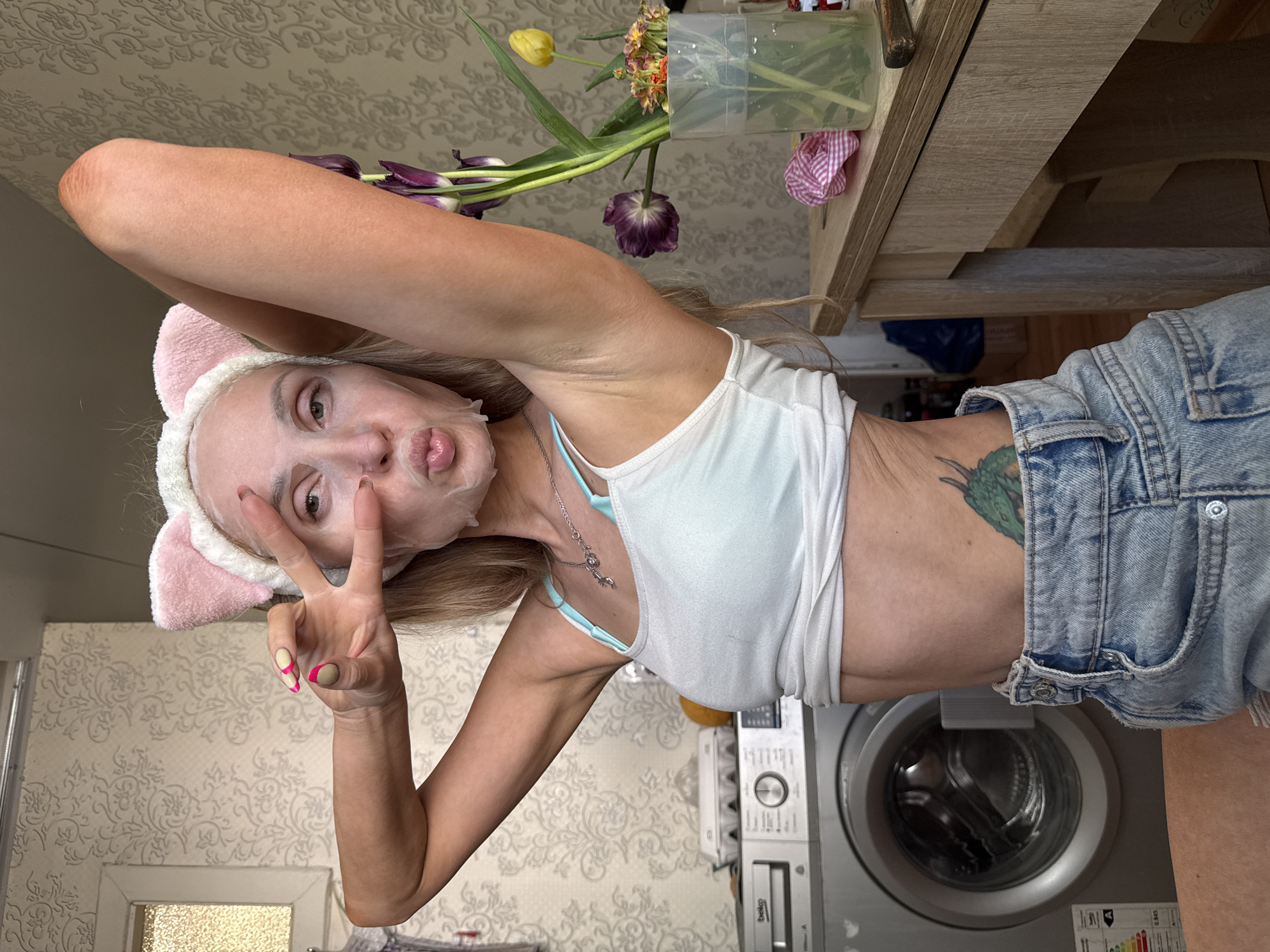A woman wearing a face mask and pink headband with bunny ears poses indoors, making a peace sign with her left hand. She is wearing a white tank top and light blue denim shorts, with a tattoo visible on her right side. Behind her is a washing machine and a table with flowers in a vase. The wallpaper has a patterned design. The image suggests a domestic setting, potentially a bathroom or laundry room. There is no verifiable evidence in the image to support the location being Chaikovsky, Russia.