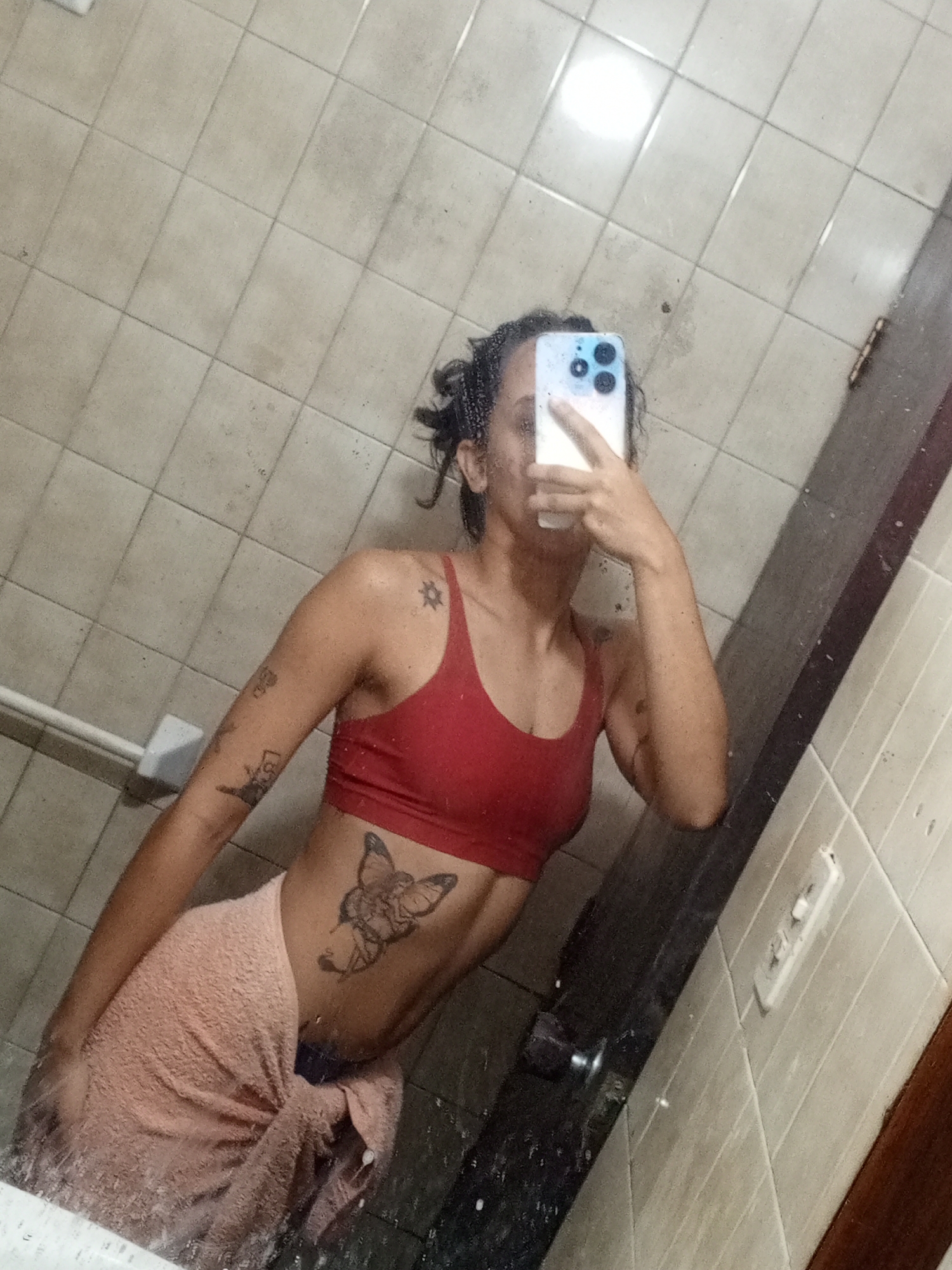 A woman is standing in a bathroom, taking a selfie in the mirror. She is wearing a red sports bra and a peach-colored towel is wrapped around her waist. The bathroom has light-colored tiled walls and a tile floor. The presence of water droplets on the mirror suggests recent use of the shower or sink. The location is a bathroom in an establishment in Porlamar, Venezuela.