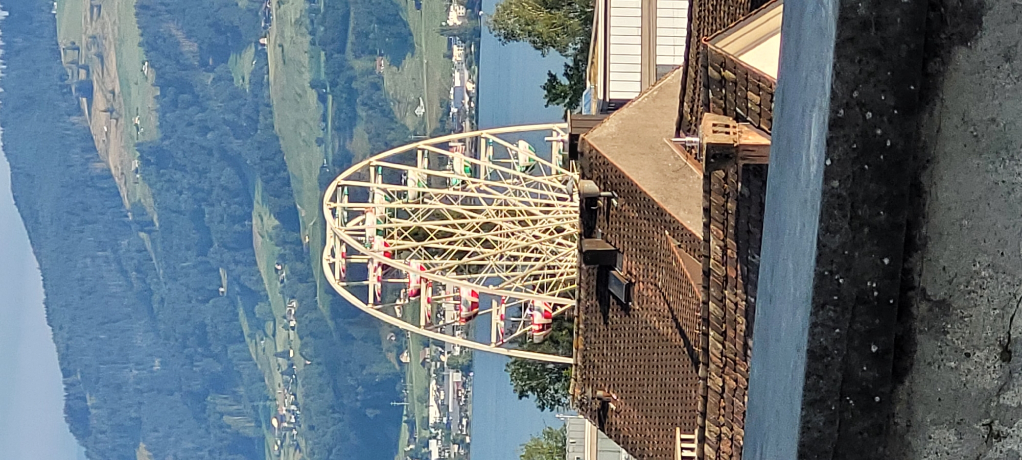 The image shows a Ferris wheel in Rapperswil-Jona, Switzerland, with a lake and hills in the background. The Ferris wheel is white with red and green gondolas. It is a sunny day, and the sky is blue. In the foreground, there are rooftops of houses. The Ferris wheel is located on the shores of Lake Zurich, and the hills in the background are part of the Swiss Alps. The city of Rapperswil-Jona is a picturesque town known for its medieval castle and charming old town. The Ferris wheel is a popular attraction for tourists and locals alike, offering panoramic views of the surrounding landscape.
