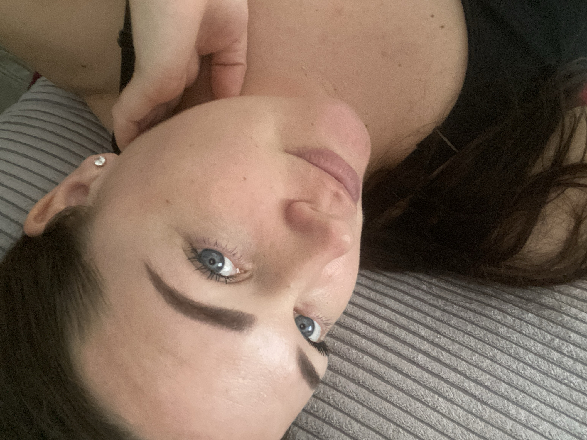 A fair-skinned woman with dark brown hair and blue eyes is lying on a grey striped surface, possibly a bed or couch. Her head is tilted towards the camera, with her right hand near her neck/shoulder area. She has dark eyebrows and visible eyelashes. A small earring is visible in her left ear. The background is blurred, but it appears she is indoors. There is no evidence to suggest that this photo was taken in Sochi, Russia.