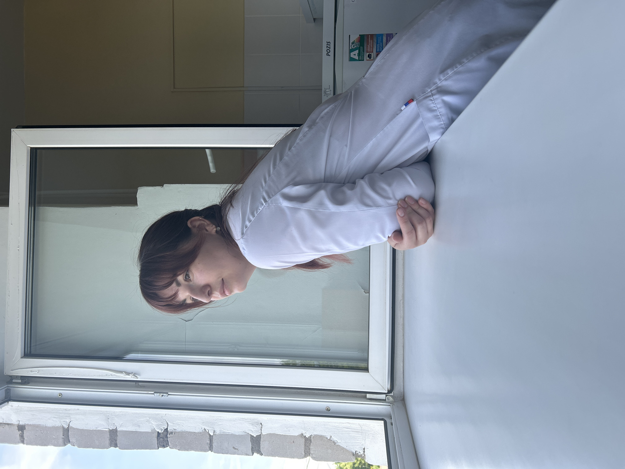 A woman with brown hair, wearing a white lab coat with a small flag on the sleeve, is looking out of an open window. The window is white-framed and overlooks a brick wall. The background shows a light-colored wall and a piece of white equipment with the word "POZIS" on it. The location is Фоки, Russia.