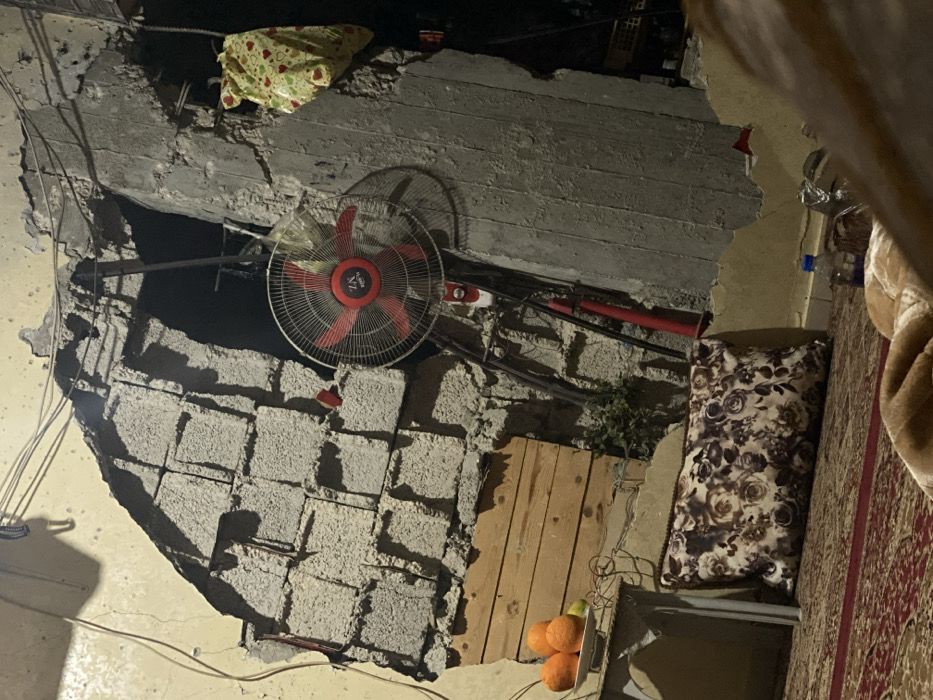 A damaged interior shows exposed concrete blocks in a ceiling. A red and silver fan is attached to the ceiling. A plastic bag rests above the fan. Wooden planks patch a section of the wall, near a cluster of oranges. A patterned pillow leans against the wall, adjacent to a patterned rug. Electrical wires are visible throughout the damaged area.