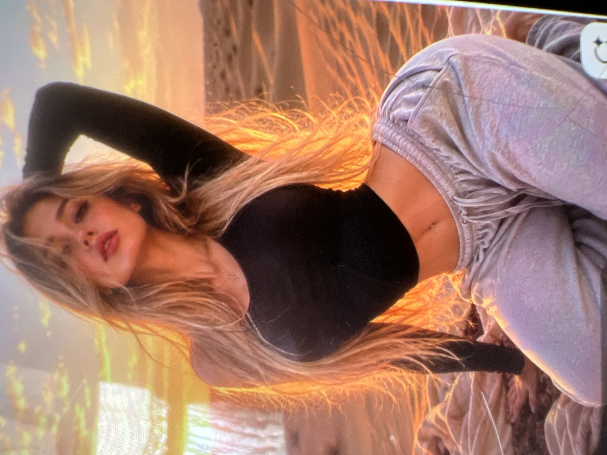 A woman with long, blonde hair is posing outdoors. She is wearing a black long-sleeved top and light grey sweatpants. The woman is kneeling on a beige blanket. Behind her is a desert landscape with sand dunes, illuminated by the warm glow of a sunset. The sky is a blend of orange, yellow, and pink hues, with some clouds visible. The scene appears to be a photoshoot.