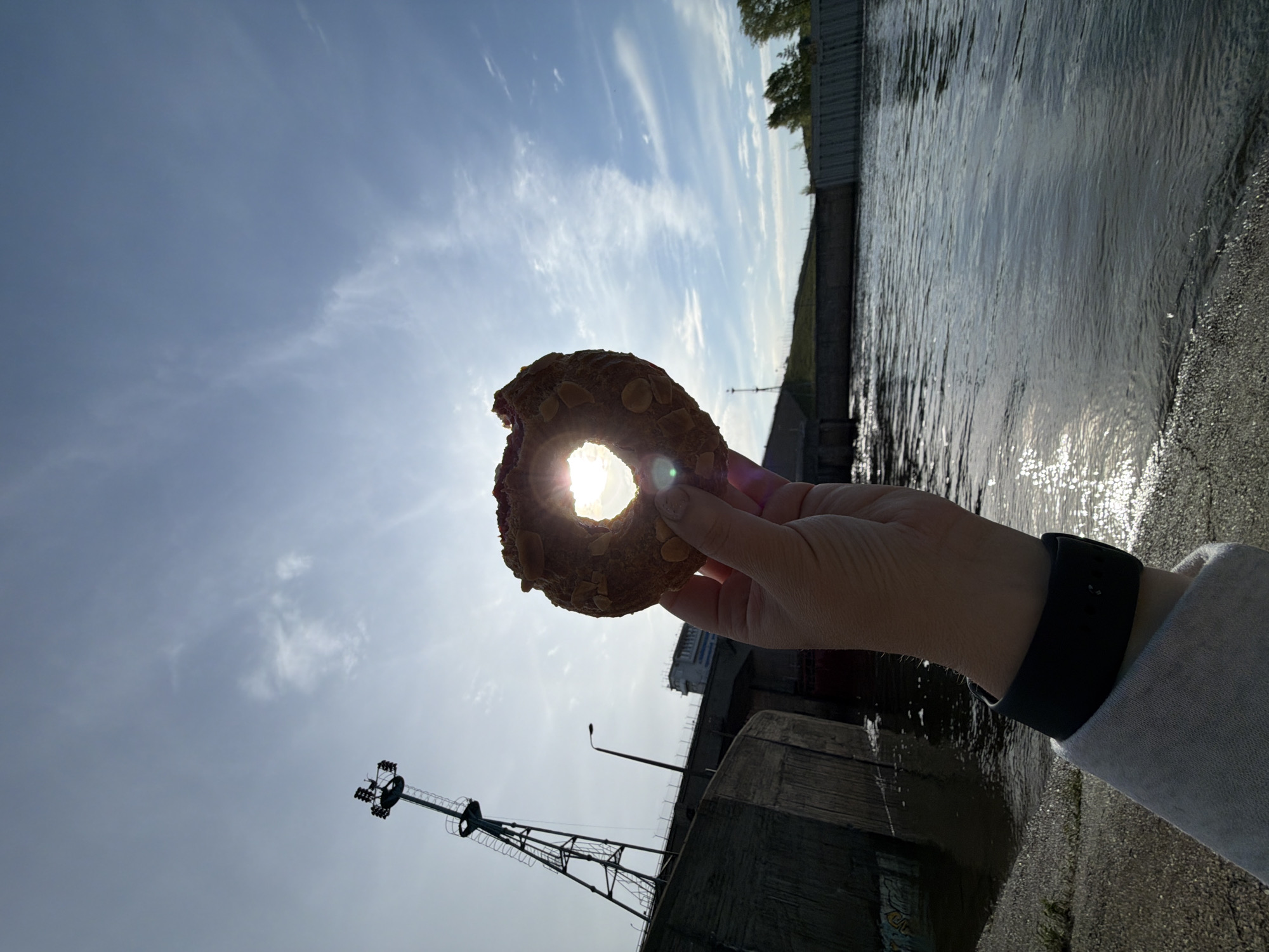 A person holds a donut in front of a bright sky, using the hole in the donut to frame the sun. The donut appears to be topped with almonds. The person is wearing a light-colored jacket and a black smartwatch. In the background, there is a body of water with some reflections, concrete structures, vegetation, a lamppost, and a crane-like structure. Based on available information, this scene seems to be located in Chaikovsky, Russia, near the waterside.