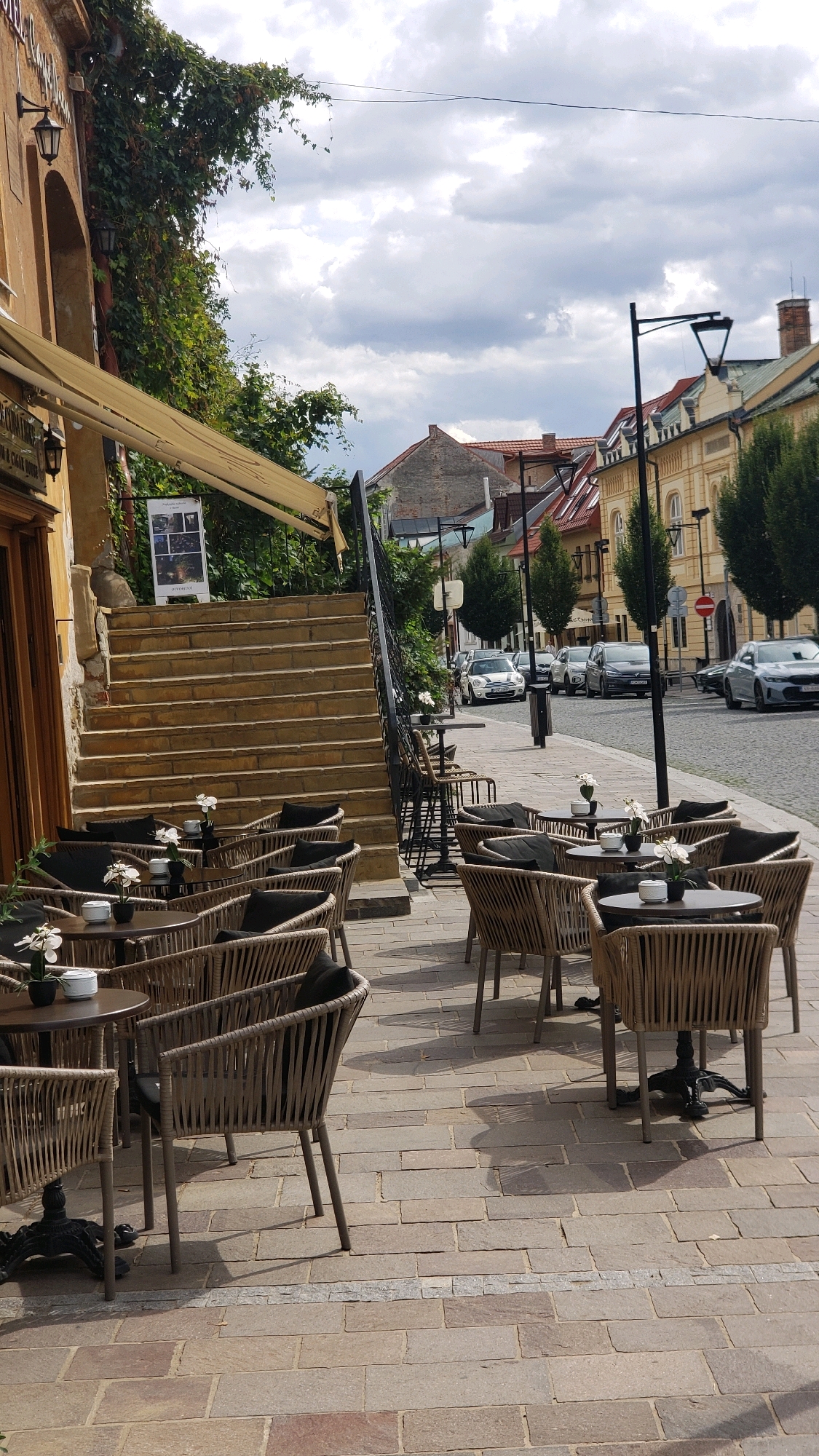 The image depicts an outdoor cafe seating area on a cobblestone street in Prešov, Slovakia. In the foreground, several round tables with chairs are arranged on a paved patio. The chairs are made of a woven material and have dark cushions. Small white flowers in black pots adorn some of the tables. Behind the cafe seating, a wide set of stone stairs leads up to an archway and a building with ivy growing on its wall. The street itself is lined with buildings and trees, and several cars are parked or driving along it. A lamppost stands on the sidewalk. The sky is partly cloudy. The overall scene suggests a pleasant day in a European city, likely a tourist or dining area.
