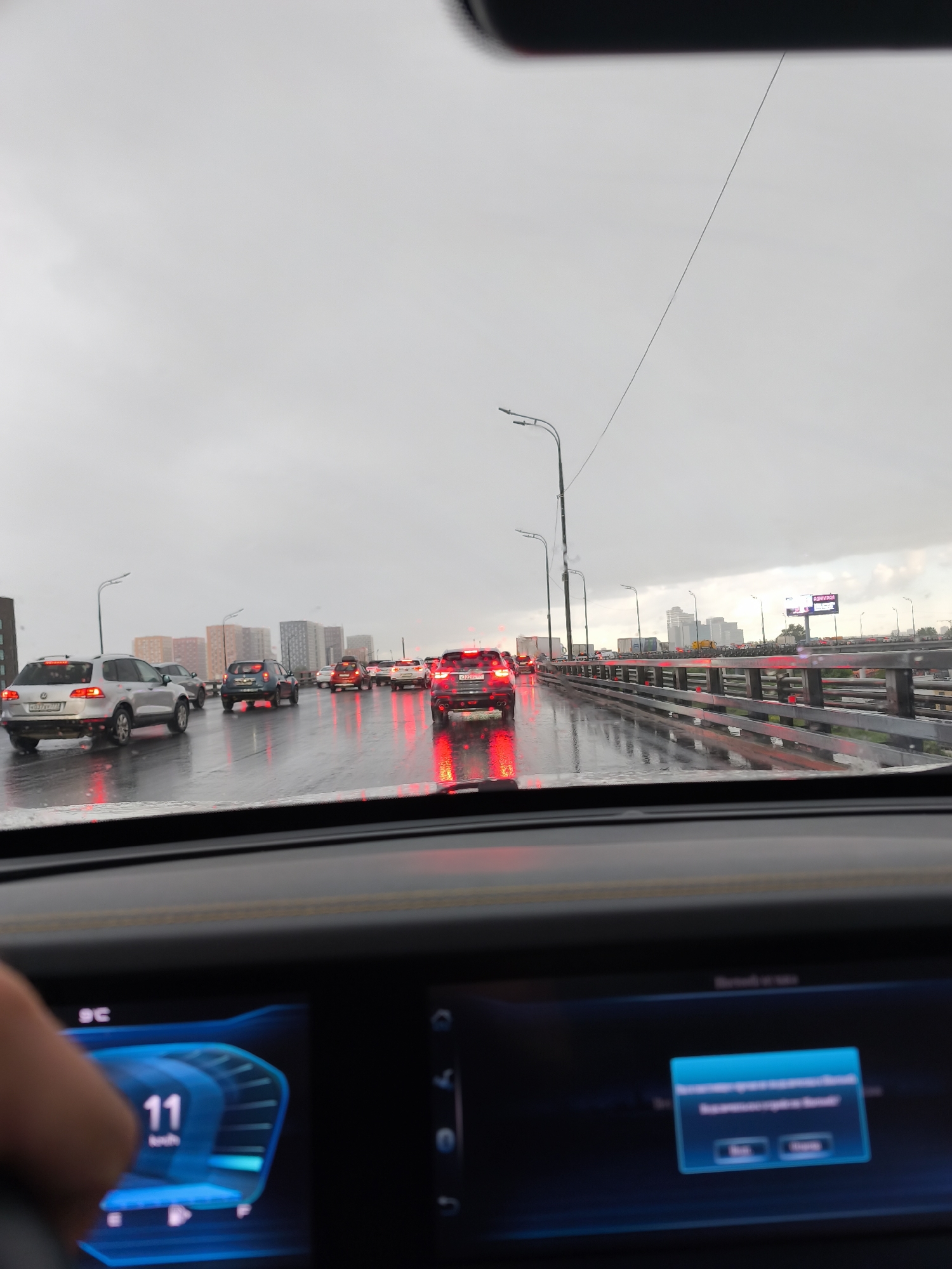 The image depicts a congested highway in Moscow, Russia, on a rainy day. The view is from inside a car, with the dashboard visible, showing a speed of 11 km/h. Several vehicles are ahead, their tail lights reflecting on the wet road. Buildings are visible in the distance, suggesting an urban area. The sky is overcast. The scene implies slow-moving traffic due to inclement weather. A billboard is visible in the distance.