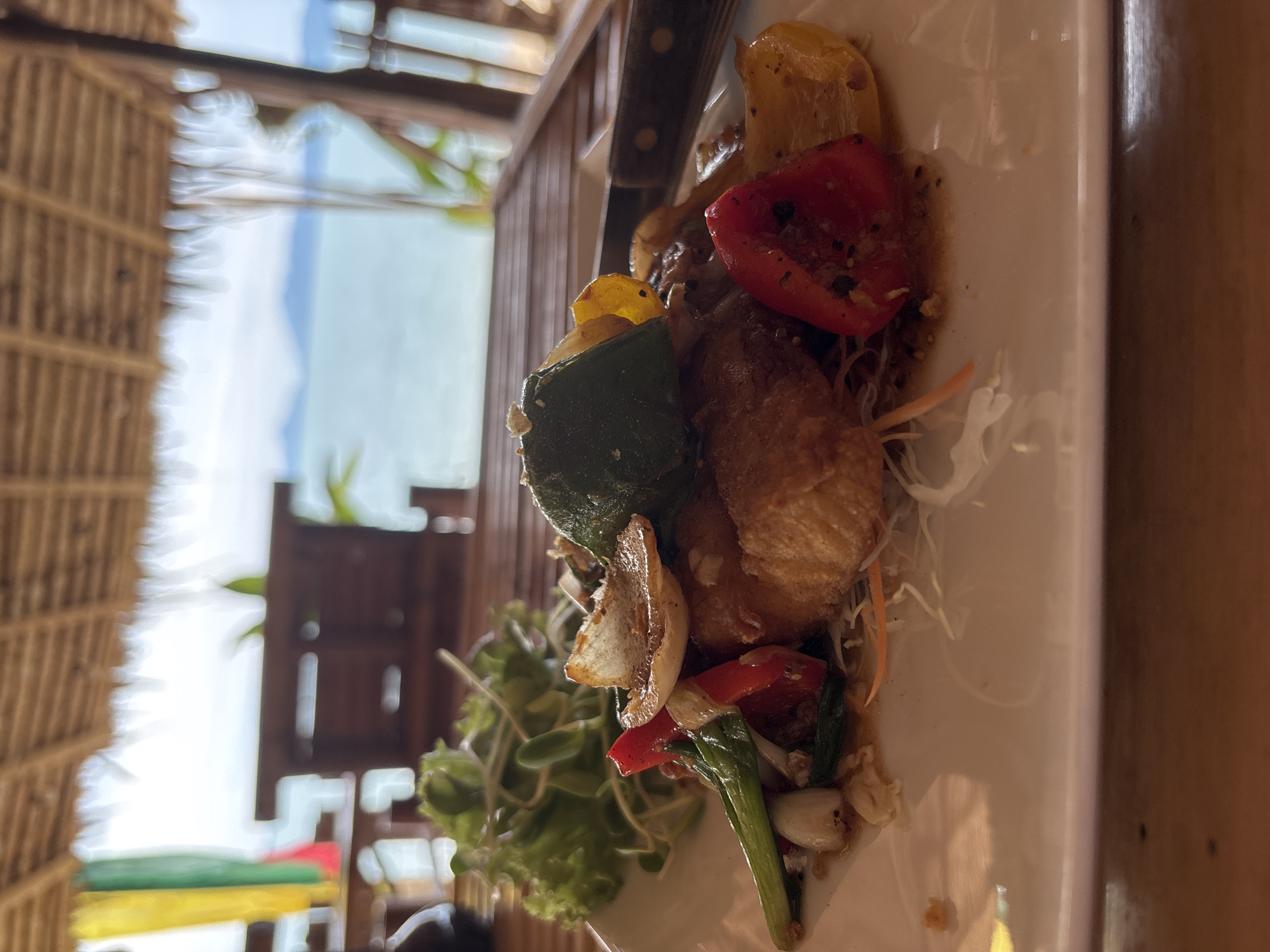 A close-up, eye-level shot shows a white plate of food on a wooden table. The food is a generous portion of what appears to be fried fish, topped with sautéed vegetables including red and yellow bell peppers, green onions, and sliced onions, garnished with a leaf of lettuce and sprouts. In the background, a thatched roof structure suggests an open-air restaurant, overlooking a body of water, possibly the sea, with islands in the distance, consistent with the coastal scenery of Baan Bo Phut, Thailand. The overall lighting is natural, coming from the outdoor environment.