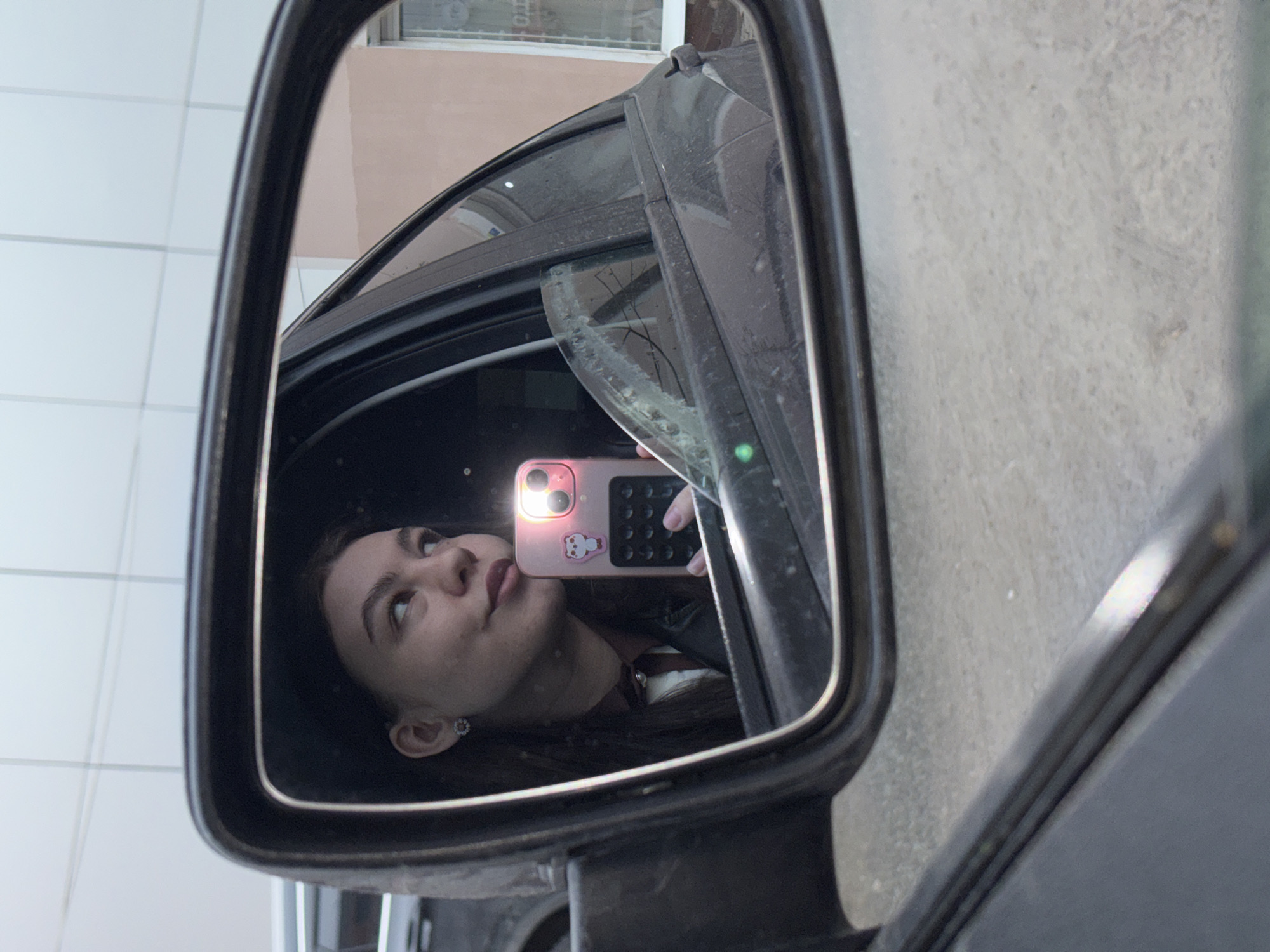 A rearview mirror on a car shows the reflection of a woman holding a pink smartphone with a built-in flash activated, reflecting in her face. The window is covered in dust. The location is near a building in the city of Perm, Russia, as evidenced by Cyrillic script visible through the window.