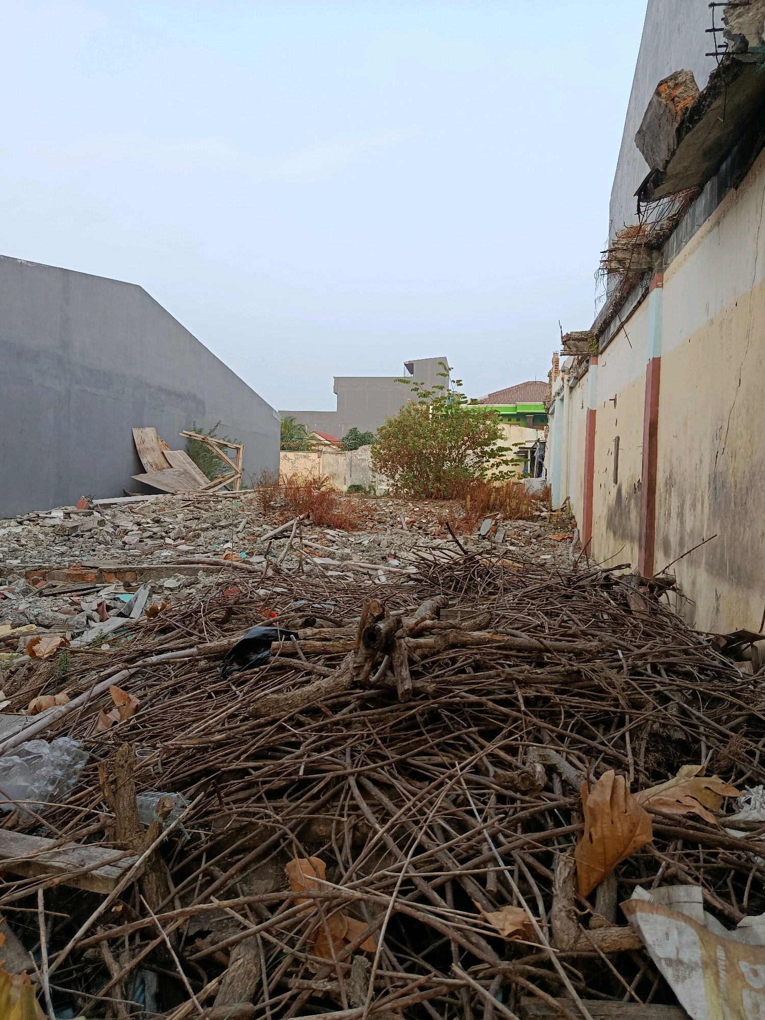 A pile of debris, consisting of branches, leaves, and construction materials, occupies the foreground of an outdoor scene in Bekasi, Indonesia. The debris is situated between two buildings. To the left is a plain gray wall. To the right is a light-colored building with a weathered facade, featuring visible cracks and faded paint. The sky above is overcast and pale. In the background, other buildings and vegetation are visible, suggesting an urban or suburban environment. The overall impression is one of disarray and abandonment.