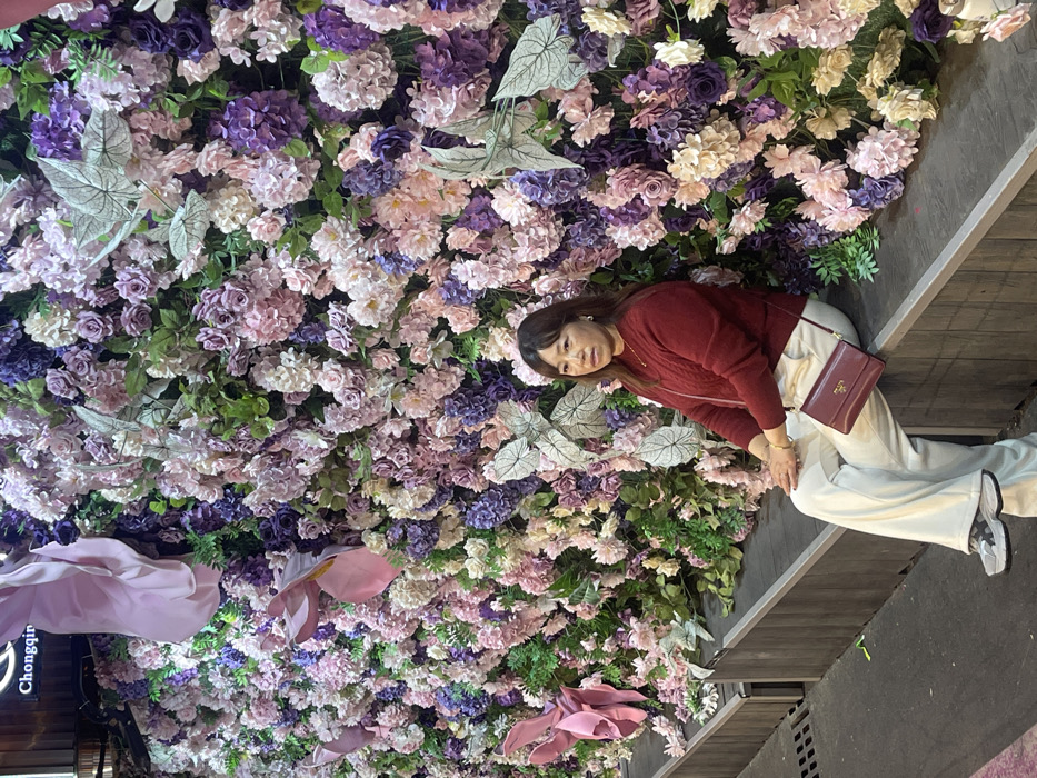 A full-body shot captures a woman posing casually in front of a vibrant, densely decorated floral wall. The image is oriented vertically, with the woman positioned towards the right side of the frame, leaning against a dark, wooden-textured surface.
The woman has shoulder-length brown hair and is looking towards the camera with a gentle smile. She is dressed in a deep red long-sleeved, possibly knitted, top. She wears light-colored, likely cream or off-white, wide-legged trousers and sneakers with light and dark gray detailing. A dark red shoulder bag, identifiable by the visible "PRADA" logo on its front flap, hangs across her body. Her left hand is resting on her lap or thigh, while her right arm is bent, holding the strap of her bag. She has small earrings and possibly a delicate necklace.
The dominant background is an elaborate wall adorned with artificial flowers and foliage. The flowers are a mix of shades, including various purples (lavender, violet), soft pinks, and creams or whites, creating a rich tapestry of blooms. Many of the flower clusters resemble hydrangeas and roses. Interspersed among the flowers are green leaves, as well as distinct, larger silver-veined leaves, adding texture and visual interest. Towards the lower left corner of the floral wall, there are also a few large, stylized, fabric flower-like decorations in a soft pink hue.
To the woman's right, a dark, horizontal surface with a wood-grain texture is visible, appearing to be a bench, ledge, or a section of a wall, against which she is leaning. In the upper left corner of the overall scene (which appears at the bottom left if the image is viewed in its original rotated orientation), a dark sign with white text is partially visible, showing the letters "Chongqin," likely part of the word "Chongqing."
The lighting in the image is soft and even, suggesting an indoor setting or a well-lit outdoor area designed for photography. The overall impression is that of a posed snapshot in a beautifully decorated environment.