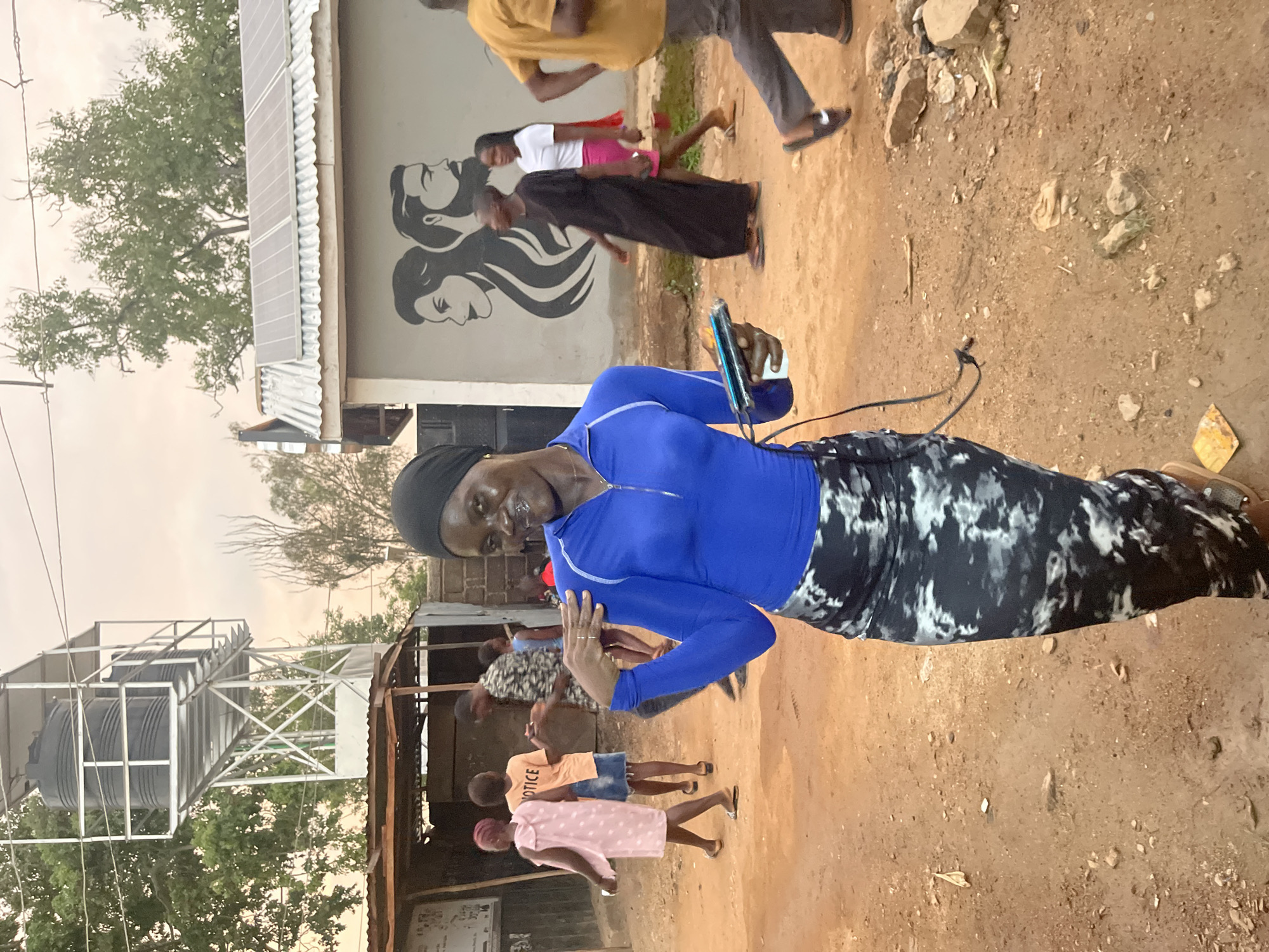 The image depicts a street scene, likely in Jos, Nigeria, with a young woman in the foreground, smiling at the camera. She is wearing a bright blue long-sleeved shirt and a patterned skirt. She holds a mobile device with cables attached in her right hand. In the background, other people are visible, including children and adults, some walking and others gathered. The environment appears to be an unpaved street, with buildings and a water tower in the distance. One building features solar panels on its roof and a mural of two profiles. The overall impression is of a community setting, possibly a residential or commercial area.