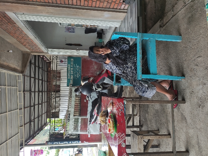 This image, captured in Kepanjen, Indonesia, portrays a street or semi-outdoor scene during the daytime. The primary subject is a woman sitting on a vibrant blue wooden chair. She wears a patterned black and white or dark blue dress or garment, with her hands clasped in front of her. Her dark hair is visible, and her gaze is directed towards the left side of the frame, suggesting a moment of rest or waiting. She appears calm, though her expression is not clearly discernible.

Beside her, a dark-colored motorcycle is parked. In front of the woman and the motorcycle is a red table, laden with various items, including what appears to be packaged food or merchandise wrapped in plastic. The setting includes a protective overhead structure made of metal framework, likely a roof or awning, indicating a sheltered area for a stall or entrance. A brick wall is visible in the upper left, contributing to the architectural context. The ground beneath is concrete or paved.

A significant detail is a green sign mounted on a wall in the background. The visible text on this sign clearly reads "BANTUAN HUKUM," which translates to "Legal Aid." Below this, another line of text reads "ADANA SATYA YULISTISI," likely the name of the legal aid organization or a person associated with it. Smaller, less distinct text is also visible beneath the main inscription. The overall scene suggests a common street-side or shop-front environment in an Indonesian town, possibly near a legal services office. The lighting is soft, implying an overcast day or that the area is in shade.