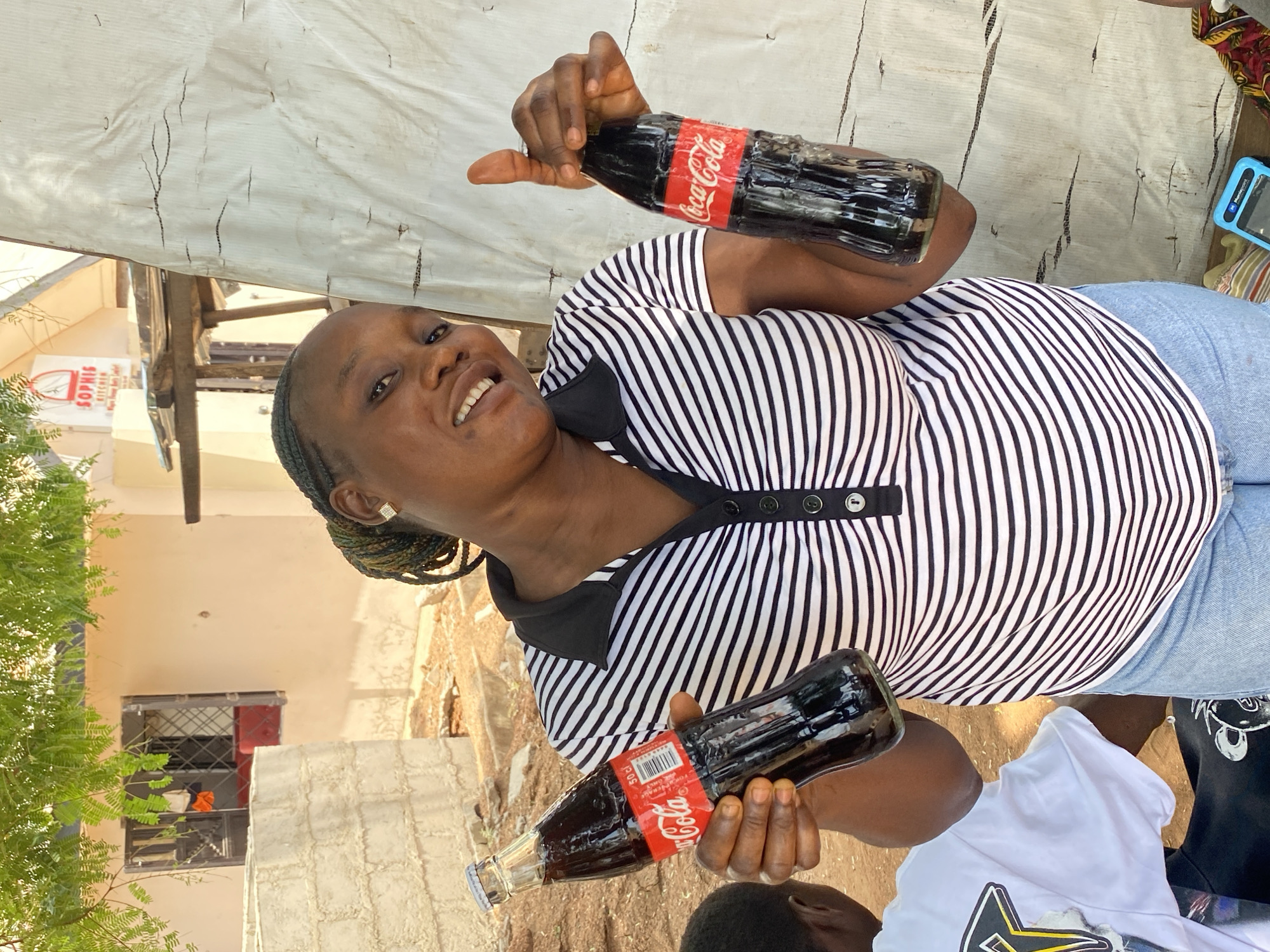 A woman with dark skin and dark braided hair stands outside in Zango-ndaji, Nigeria, holding two glass bottles of Coca-Cola. She is wearing a black and white striped shirt, blue jeans, and earrings. A tarp is seen in the background. The environment includes a building with a sign that appears to say "Sophie's Kitchen," trees, and the ground is dirt. Another person is partially seen to her right.
