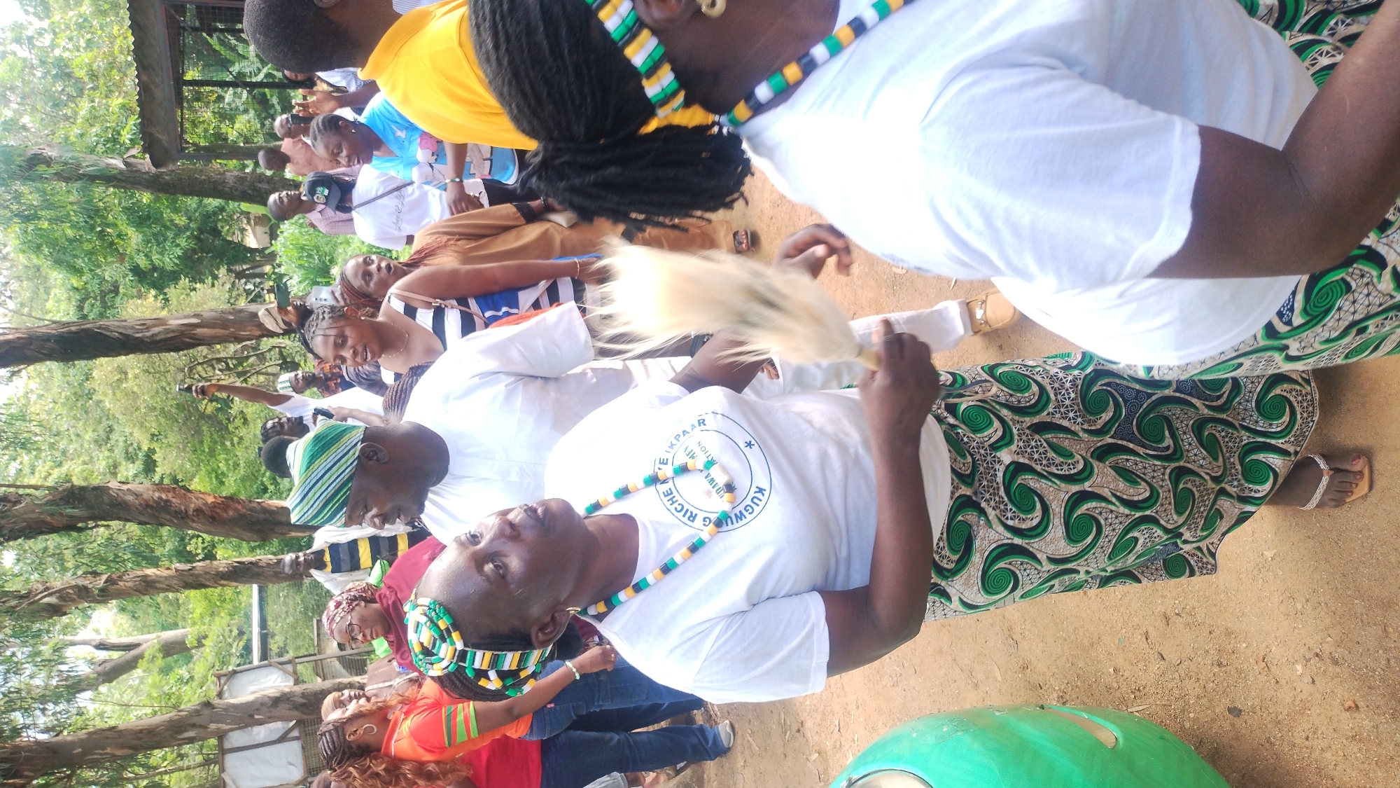 In a medium shot, an outdoor gathering is captured on a dirt path surrounded by trees and foliage, likely in Jos, Nigeria. The scene focuses on several individuals, predominantly women, dressed in vibrant clothing and traditional attire. In the foreground, a woman wearing a white t-shirt with a circular logo and a green, black, and white patterned skirt is holding a fluffy white object. She also wears a matching beaded necklace and headband. To her left, another woman in a colorful orange top and blue jeans is visible. A man in a white shirt and a green and white striped cap is positioned behind the woman with the white t-shirt. In the background, a diverse crowd of people, both men and women, are engaged in conversation and observation, with some holding mobile phones. The atmosphere appears festive or celebratory.