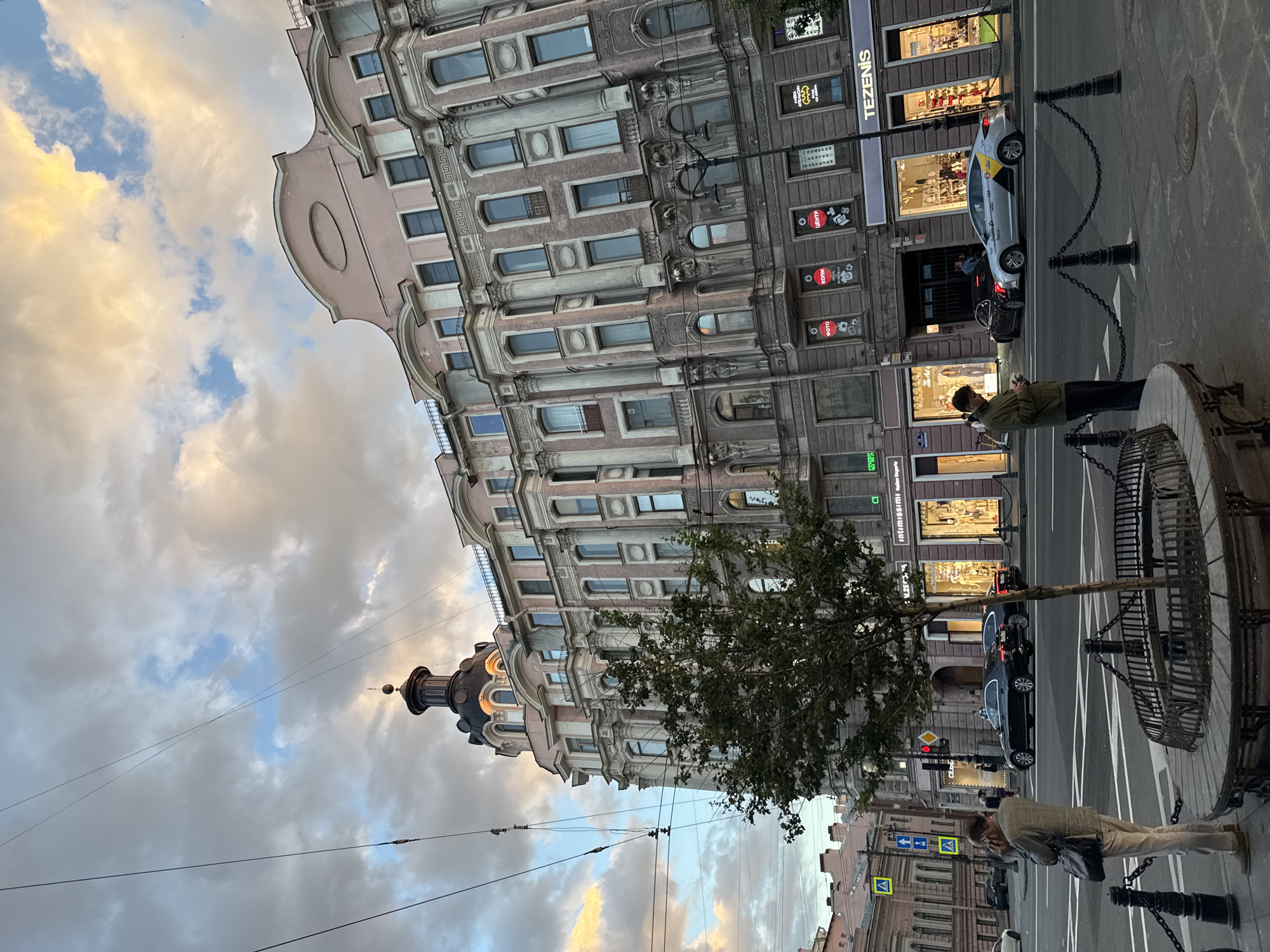 The image depicts a street view in Saint Petersburg, Russia, with a grand, ornate building on the right side of the frame. The building exhibits classical architectural features, including multiple stories, numerous windows of varying shapes and sizes, decorative columns, and a rounded turret topped with a spire. The sky above is filled with dramatic clouds, illuminated by the warm glow of sunset, creating a striking contrast with the dusky tones of the city.
In the foreground, a young man stands on the sidewalk, looking down at his phone. He is wearing a green jacket and dark pants. To his left, a woman is seen from behind, wearing a light-colored jacket and pants, and carrying a dark shoulder bag. She is also looking towards the street.
The street is paved and appears to be an intersection. Several cars are visible, including a black sedan and a silver taxi with a yellow and black stripe. A small tree, enclosed by a decorative metal fence, stands in the middle of the sidewalk. In the background, more buildings line the street, and traffic signals and street signs are present. Shops with illuminated storefronts are visible on the ground floor of the main building, with signs such as "TEZENIS" and others in Cyrillic script. The overall scene captures a moment in time on a city street, with a blend of architectural grandeur and everyday life under a beautiful, cloud-filled sky.