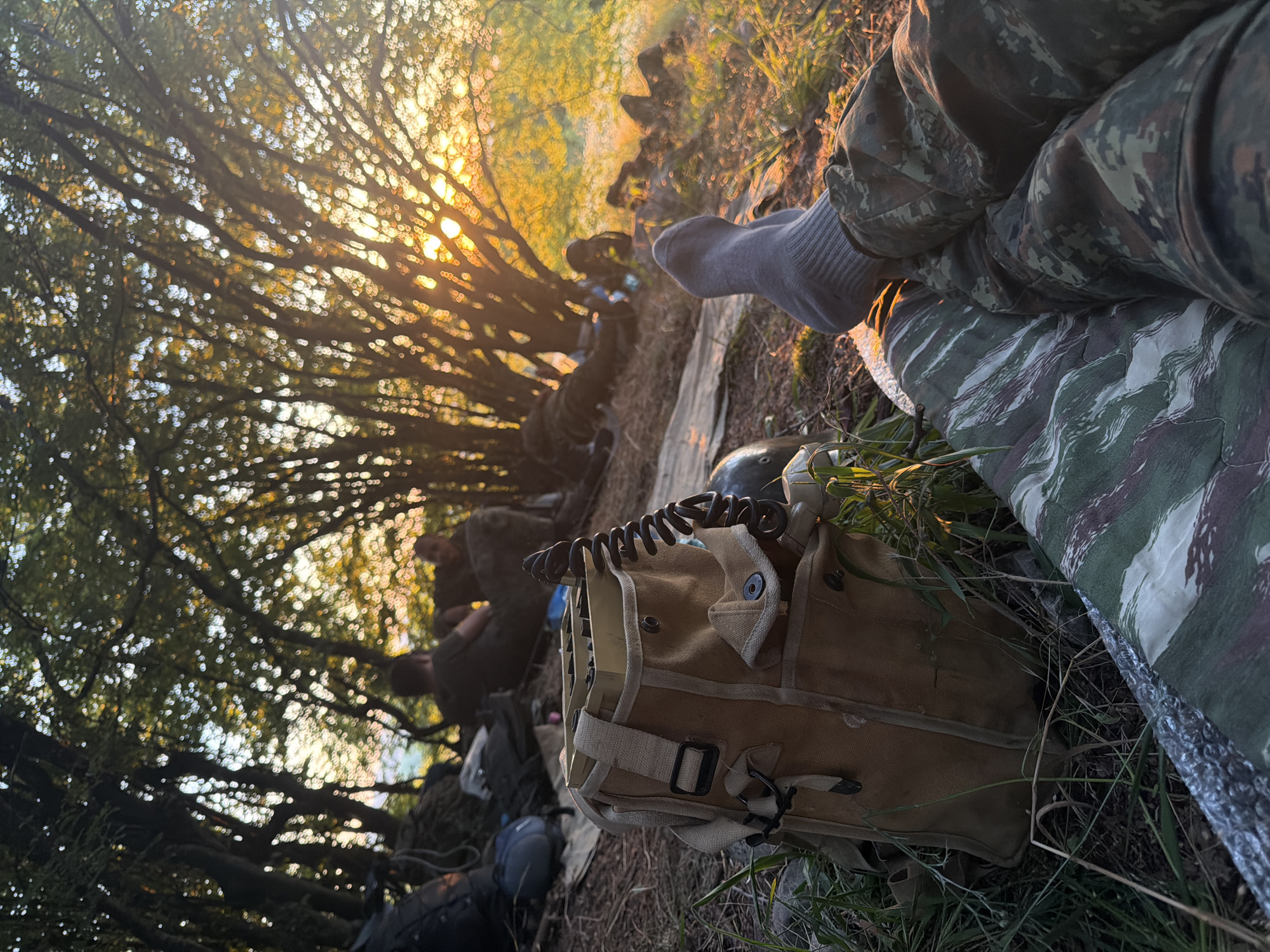 This media file depicts a group of people in military attire resting outdoors as the sun sets. The setting appears to be a wooded area, with dappled sunlight filtering through the trees, creating a warm, golden glow. The people are lying down, some with their heads on bags, suggesting a break or encampment.
The primary objects visible include military-style bags, some made of canvas and others of a dark material. A khaki-colored pouch with straps and buckles is prominently featured in the foreground, with a coiled black cord extending from it. One person's legs are visible, clad in camouflage trousers and gray socks, with a camouflage-patterned blanket or mat beside them.
The time of day is clearly evening, given the low angle of the sun and the warm, diffused light. The weather appears to be pleasant, with no indication of rain or harsh conditions. The location, Ledenik, Bulgaria, provides a geographical context for this outdoor scene. While no specific emotions are explicitly conveyed, the relaxed postures and the peaceful lighting suggest a moment of respite. There is no visible text in the image. The overall impression is one of a military group taking a break during an outdoor exercise or deployment as the day concludes.