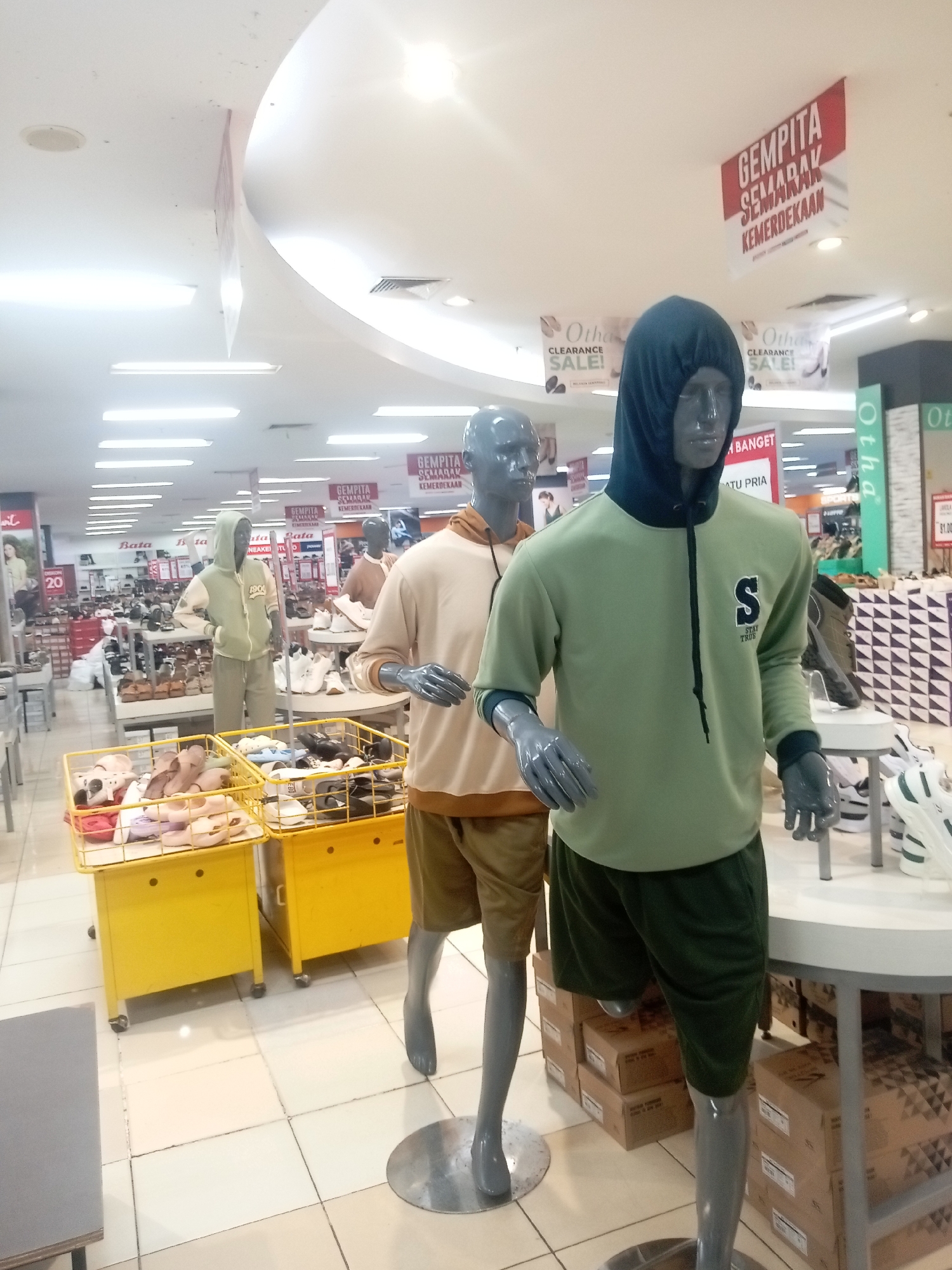 The image depicts the interior of a retail store, likely a shoe or apparel store, in Batam, Indonesia. The scene is illuminated by fluorescent lighting, casting a bright, even glow. In the foreground, two mannequins are prominently displayed. The mannequin on the right is dressed in a green hooded sweatshirt with a dark blue hood and trim, and dark green shorts. It has the letter "S" and the text "STAY TRUE" on the front of the sweatshirt. The mannequin on the left is wearing a light brown hooded sweatshirt and light brown shorts. Both mannequins have gray skin and are positioned as if walking.
Behind the mannequins, several rows of shelves are visible, stocked with footwear, including sandals and athletic shoes. Two bright yellow display carts filled with footwear are positioned in the mid-ground. Various signs are visible throughout the store, including a large red and white banner at the top with text in Indonesian, "GEMPITA SEMARAK KEMERDEKAAN," which translates to "Festive Celebration of Independence." Other smaller signs, such as "Oth" and "CLEARANCE SALE!", suggest ongoing promotions. In the background, more merchandise and store fixtures are visible, extending deeper into the store. The floor is tiled with white square tiles. The overall impression is that of a well-lit, busy retail environment during a promotional period.