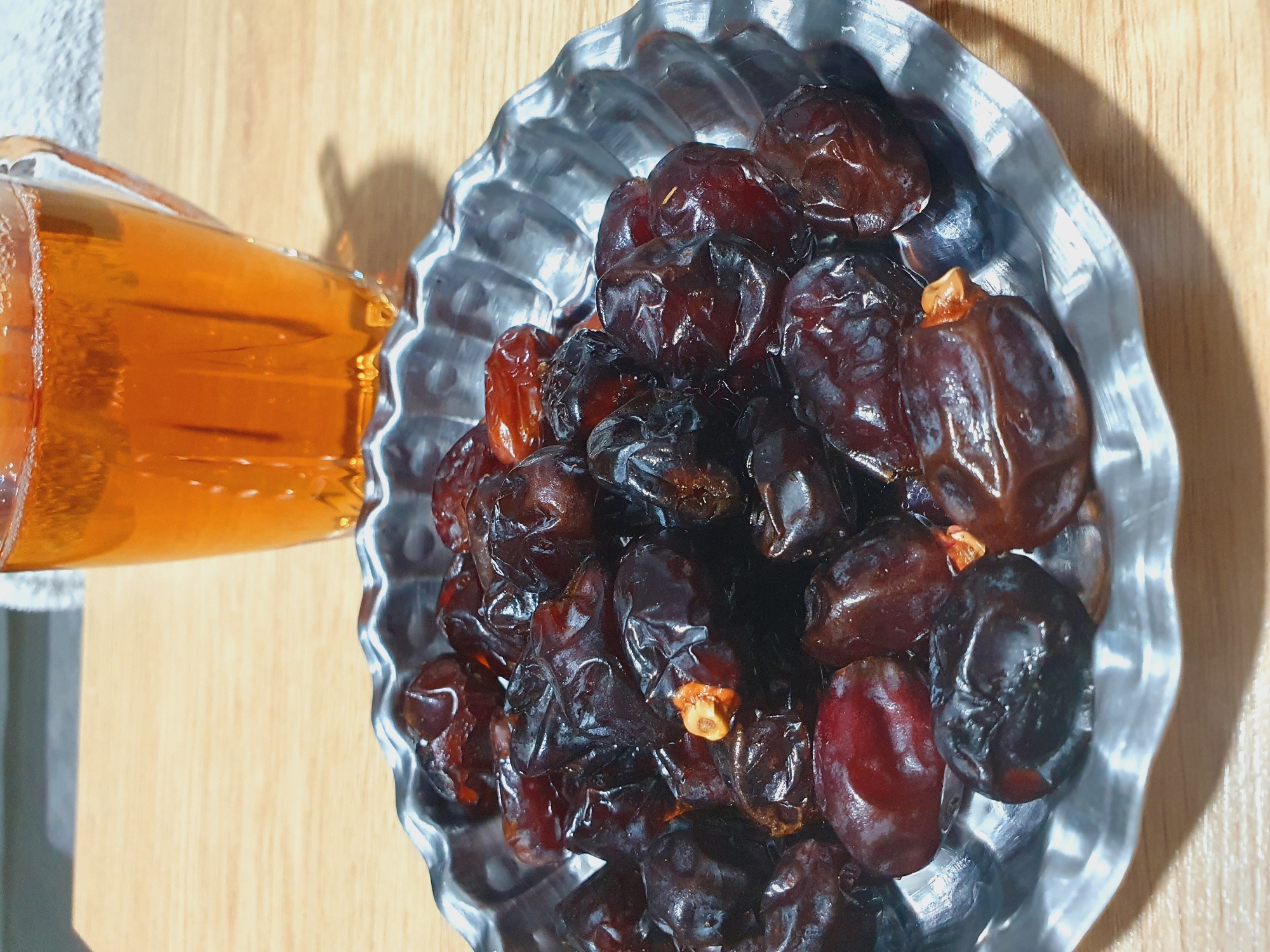 A close-up, overhead shot shows a metallic, fluted dish overflowing with dark, plump dates. The dates are a deep, rich brown, some with a reddish hue, and appear glistening, suggesting freshness or a light coating of moisture. A few of the dates reveal small, lighter-colored remnants of their pits. Behind the dish, a glass mug filled with a golden-amber liquid, likely tea, is visible. The mug has vertical ridges and a slight foamy surface at the top. Both the dish and mug rest on a light-colored wooden surface, likely a table. The background is softly blurred, with hints of a light-colored fabric or surface and a subtle shadow cast by the mug. The scene is lit, creating reflections on the dates and the mug. The image is likely a still life, capturing a common refreshment. There is no indication of location in Kheyrabad, Iran, or any people or actions taking place.