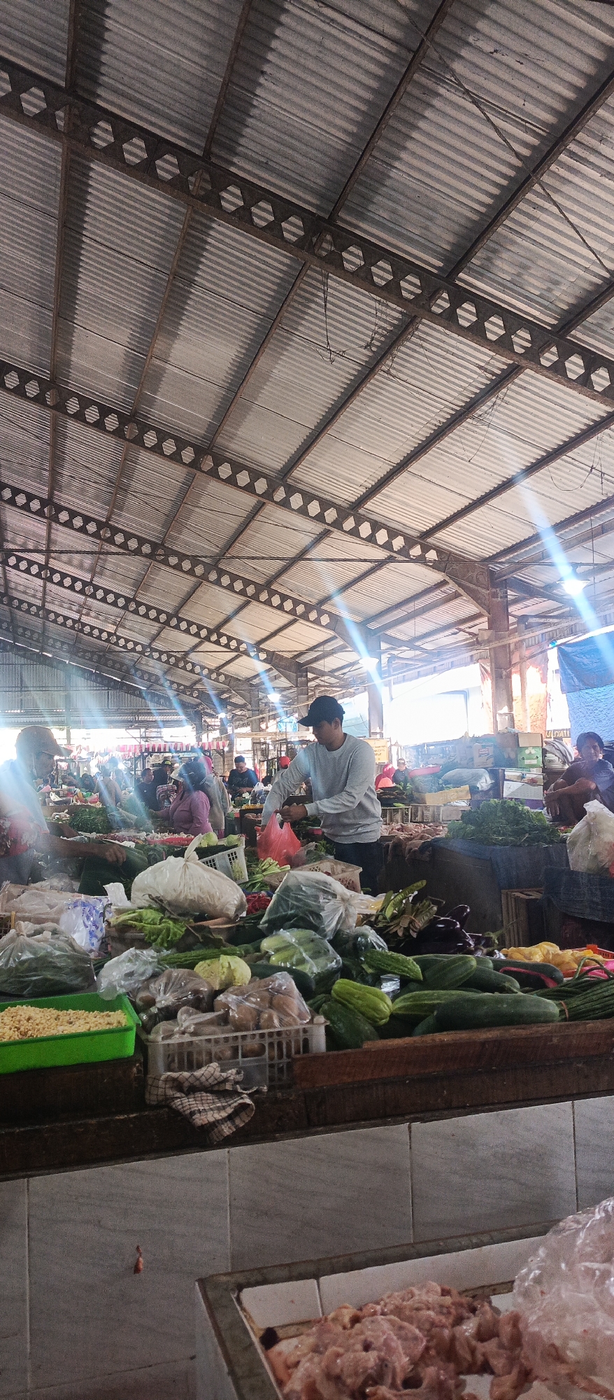 This image depicts an indoor market scene, likely a traditional wet market, possibly located in Malang, Indonesia. The structure features a corrugated metal roof supported by metal beams, with natural light entering from the sides, creating visible sun flares.
In the foreground, a wooden counter displays a variety of fresh produce, including cucumbers, eggplants, and leafy greens, some of which are packaged in plastic bags or contained within plastic crates. A bright green plastic container filled with what appears to be corn kernels is also visible.
Further into the market, several individuals are engaged in activities related to the sale of goods. A man wearing a light grey long-sleeved shirt and a dark baseball cap is seen behind a stall, holding a red plastic bag and interacting with produce. To his left, a woman in a pink garment is also attending to goods. Other shoppers and vendors are dispersed throughout the market, though their interactions are not clearly discernible. The overall environment suggests a bustling marketplace with a focus on fresh food sales. The lower part of the image shows a tiled surface and a tray filled with raw chicken pieces.