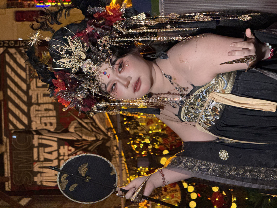 The image displays a posed, waist-up portrait of an individual in elaborate traditional-inspired attire at night. The subject is a person, likely female, with fair skin, a neutral facial expression, and intricate makeup including red eyeshadow and a red forehead embellishment.
She wears a dark, possibly black, dress or top with ornate gold and multicolored embroidery across the bodice and a visible light-colored sash. Her shoulders and arms bear subtle golden glitter. An extensive, multi-layered headpiece adorns her head, incorporating dark feathers, red floral elements, gold filigree, chains, and a prominent golden butterfly or phoenix-like motif at its center. A star-shaped ornament is visible on the right side of the headpiece. She wears multiple necklaces. On her left wrist is a red beaded bracelet, and on her right, a red and white beaded bracelet. Her right hand, held open with fingers slightly curled, is fitted with elongated, pointed gold-colored nail covers. In her left hand, she holds a circular black fan with golden cloud patterns and a dark handle.
The background is dark and blurred, illuminated by numerous warm, yellow-orange lights, indicative of an urban evening or night setting. Partially visible behind the subject's left shoulder is a banner or sign with the white English letters "SMC" prominently displayed, below which are Chinese characters "机动都市" (Jīdòng Dūshì), translating to "Mobile City" or "Mech City." This signage suggests the scene is located in a commercial or entertainment zone within Yuzhong District, China.