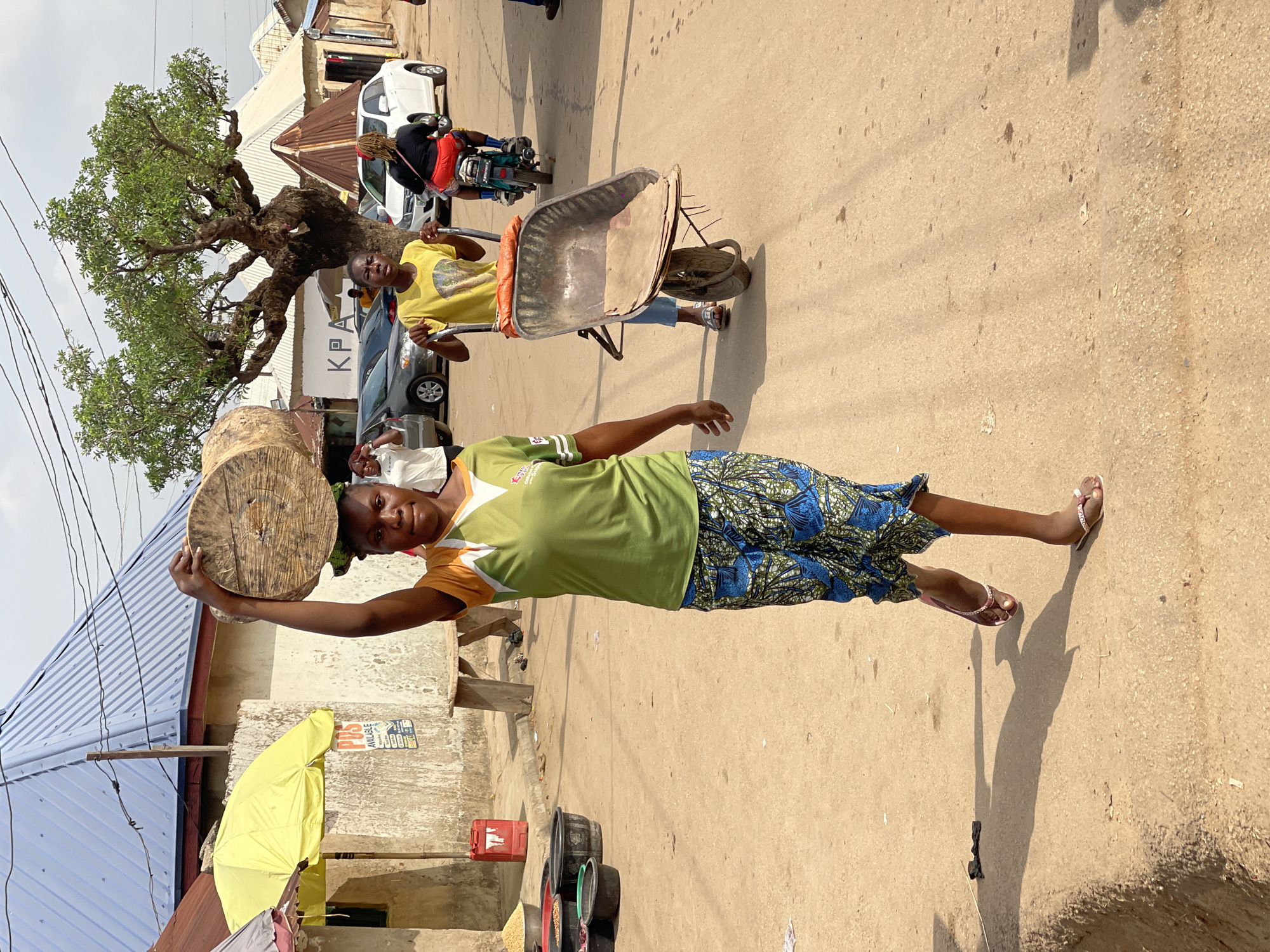 This image captures a vibrant street scene in Kuchingoro, Nigeria, under what appears to be bright daylight with some atmospheric haze.
In the foreground, a woman stands prominently, looking directly at the viewer with a slight smile. She is dressed in a light green t-shirt with orange trim and a long, patterned skirt featuring blue and black designs. Expertly balanced on her head is a large, circular wooden disc, which she steadies with her right hand.
Further down the street, a man in a yellow t-shirt pushes an empty metal wheelbarrow, also seeming to glance towards the camera. Behind him, a person on a motorcycle is partially visible, along with parked cars. Another individual wearing a white face mask can be seen behind the wheelbarrow.
The setting is a dusty, unpaved road lined with buildings, many topped with corrugated metal roofs. A large, old tree with a dense canopy of green leaves and gnarled branches is a significant feature in the mid-ground. Power lines traverse the upper left portion of the sky. Near the foreground woman, several dark plastic buckets, some holding what appear to be grains or foodstuffs, are placed on the ground alongside a red plastic container and a bright yellow umbrella.
Distinct shadows on the ground indicate a bright, sunny day, likely midday or early afternoon. Visible text includes "KPA" on a white sign behind the wheelbarrow and "POS AVAILABLE" on a small sign attached to a building near the yellow umbrella. The scene depicts everyday activity in a bustling community.