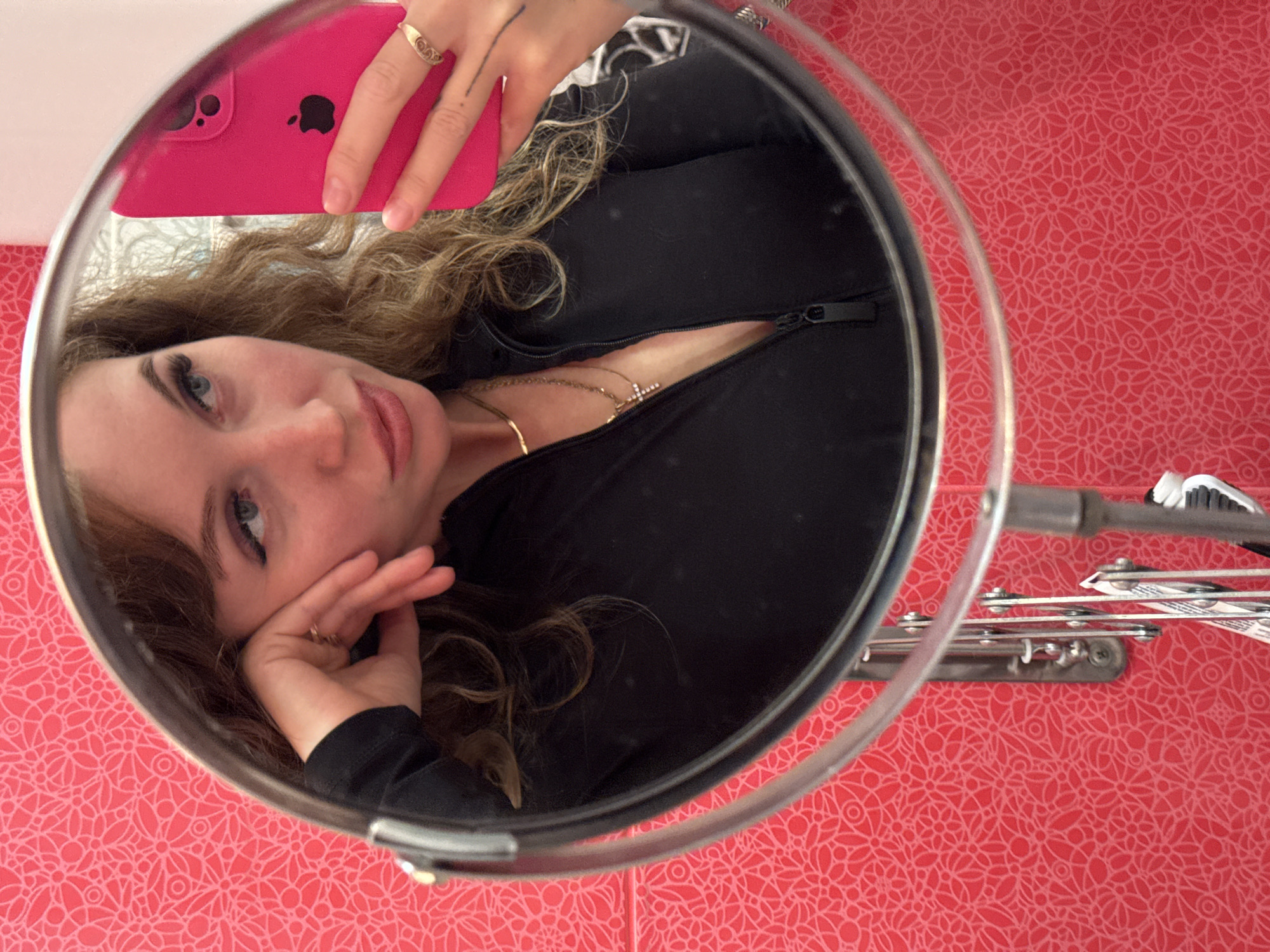 A woman is reflected in a round mirror, holding a pink smartphone. She is wearing a black zip-up jacket and has blonde, curly hair. She is looking at the camera and has a ring on her finger. A drying rack is visible in the background, which has pink floral wallpaper. It's impossible to infer from the image that the location is in the city of Chaikovsky, Russia.