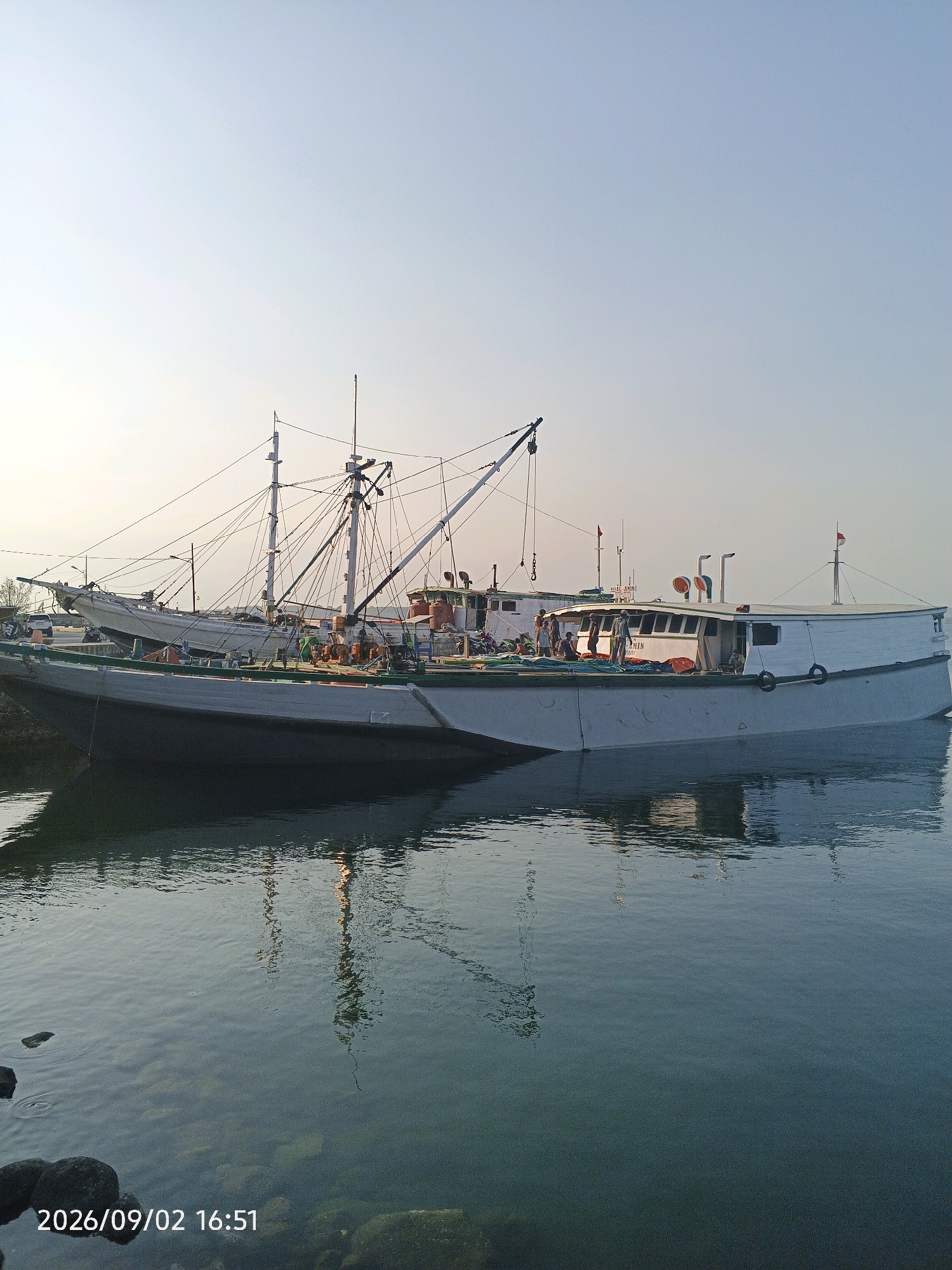 A wide-angle, eye-level shot captures two fishing boats docked at what appears to be a harbor in Pare-Pare, Indonesia, based on the embedded timestamp. The sky is clear and pale, transitioning to a light orange hue near the horizon, suggesting late afternoon or early morning. The water in the foreground is calm, reflecting the boats and the sky. The larger boat, a white vessel with a dark hull and green trim, dominates the scene. It has several masts and rigging, with some people visible on its deck. A second, smaller white boat is partially visible behind it. The overall impression is one of a working harbor at rest.