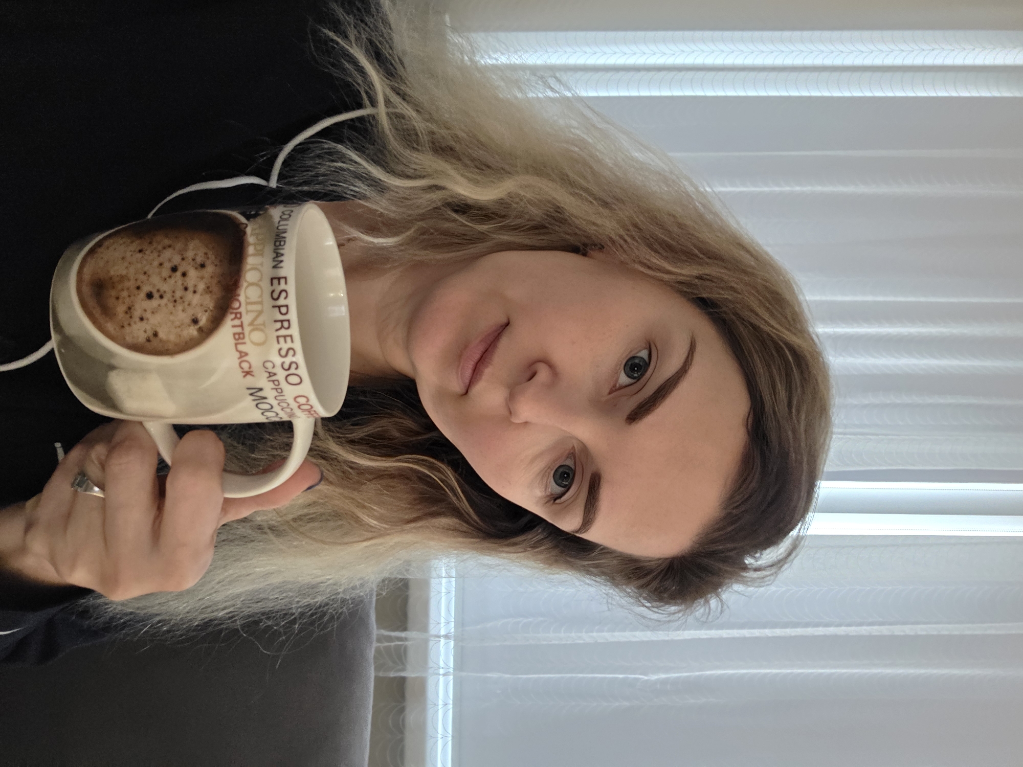 A fair-skinned woman with light brown hair holds a mug filled with coffee. The mug has the words "COLUMBIAN ESPRESSO CAPPUCCINO CORTBLACK MOCHA" printed on its side. The woman is indoors, likely near a window with white blinds. Her clothing appears to be dark-colored. She is wearing a ring on her left ring finger. There is no evidence to support the photo's location being in the city of Chaikovsky, Russia.