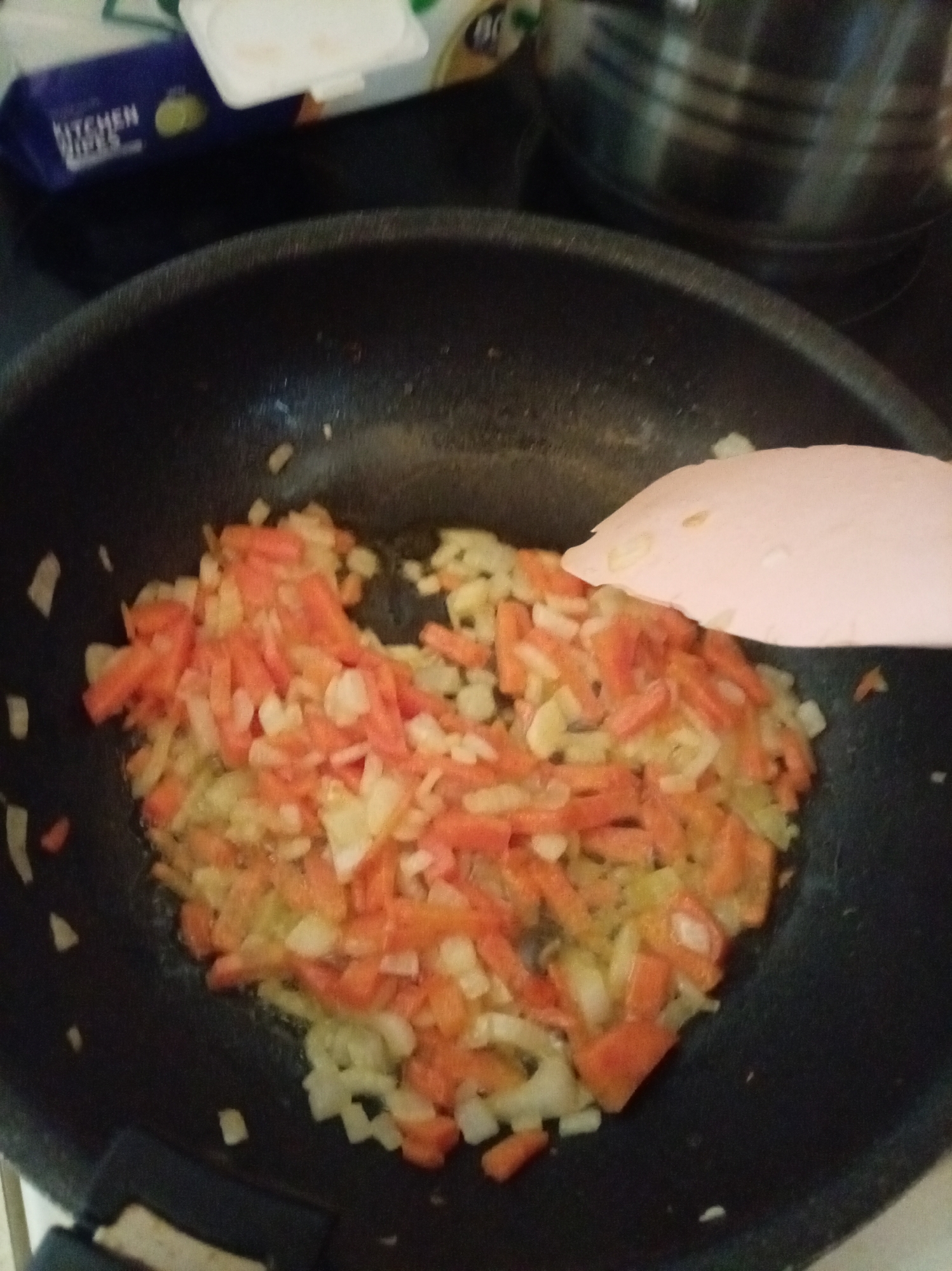 In a black frying pan, chopped carrots and onions are being sauteed. A pink spatula is partially visible, resting on top of the vegetables. The scene is likely a kitchen, with a blurred background that includes a stainless steel pot and packaging for "KITCHEN WIPES." The image is framed from a top-down perspective. This image is not of Tomtor, Russia.