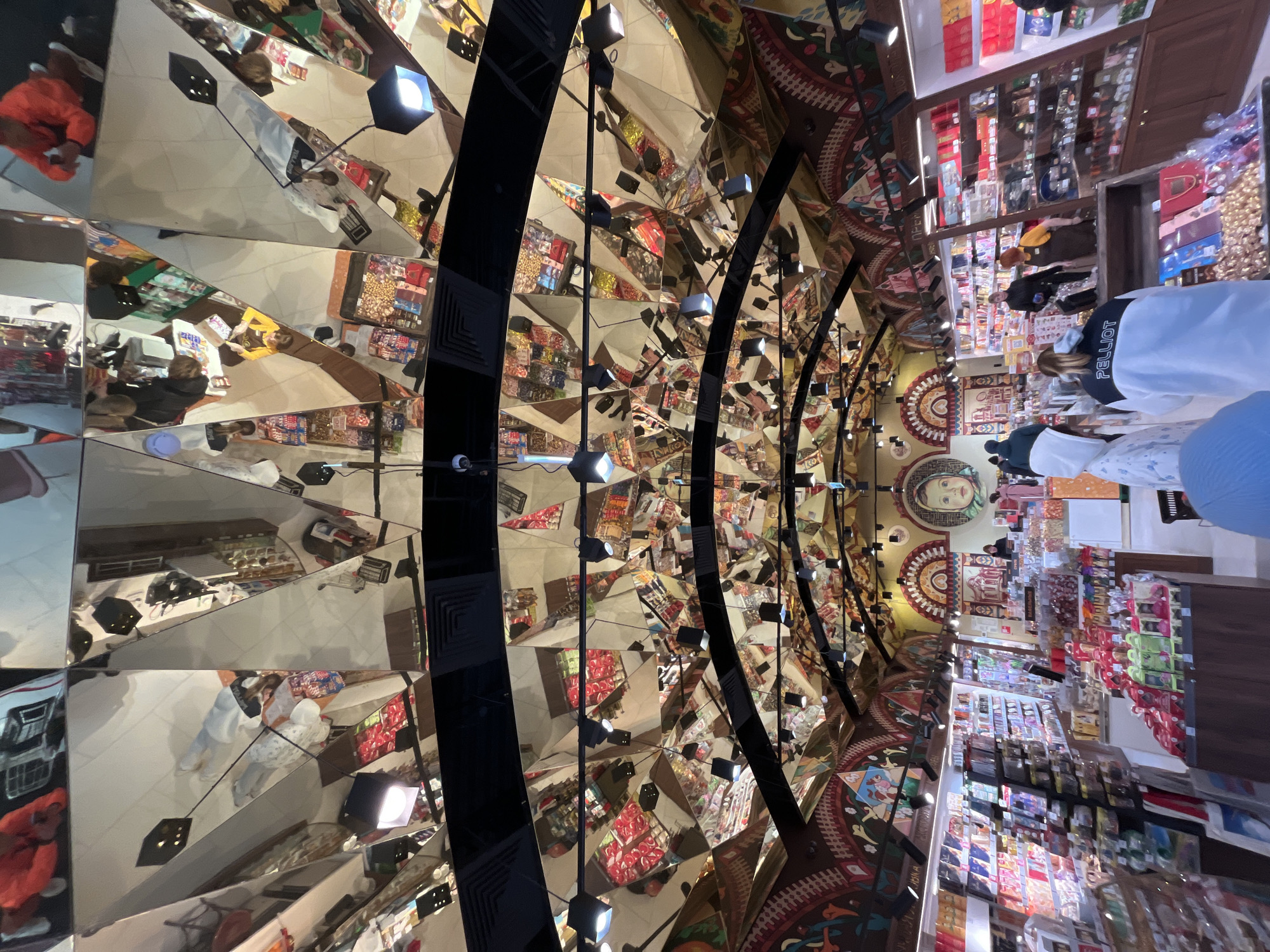 The image shows the interior of a store in Moscow, Russia. The ceiling is decorated with colorful mosaics and traditional Russian designs. Multiple mirrors reflect the store's shelves and customers, creating a kaleidoscopic effect. Black beams with spotlights are suspended from the ceiling. The store sells various food and souvenir items. People can be seen browsing the products on display.