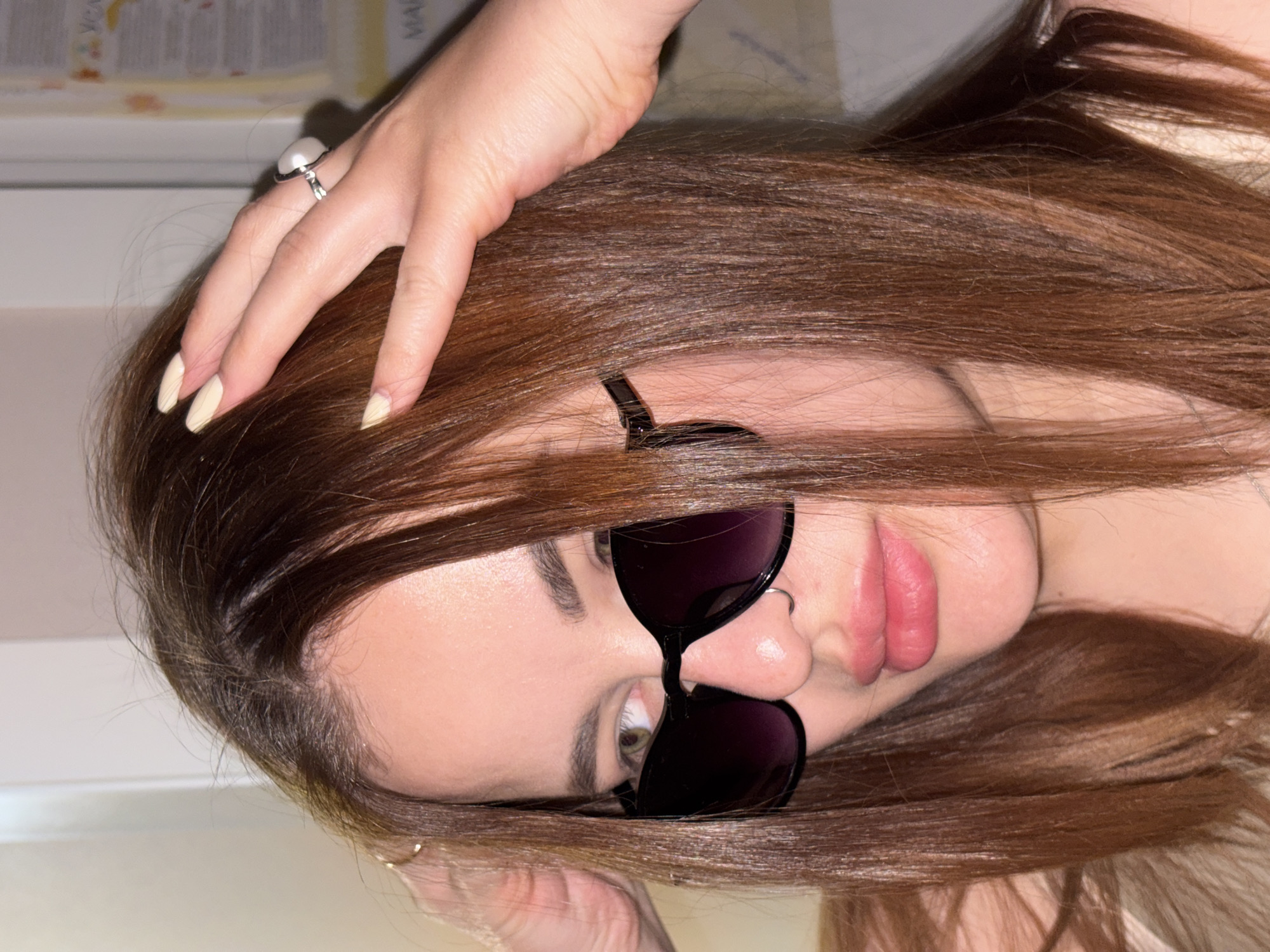 A woman with long, straight, auburn hair lies sideways with her head tilted toward the viewer. She is wearing sunglasses with dark lenses and a thin black frame. Her left hand is raised, fingers spread, with a silver ring with a pearl-like stone visible on her ring finger. The nails are painted in a light, neutral shade. A nose ring is visible in her left nostril. The background appears to be a room with a document partially visible, suggesting an indoor setting. The image does not provide information that identifies the location as Krasnodar, Russia.