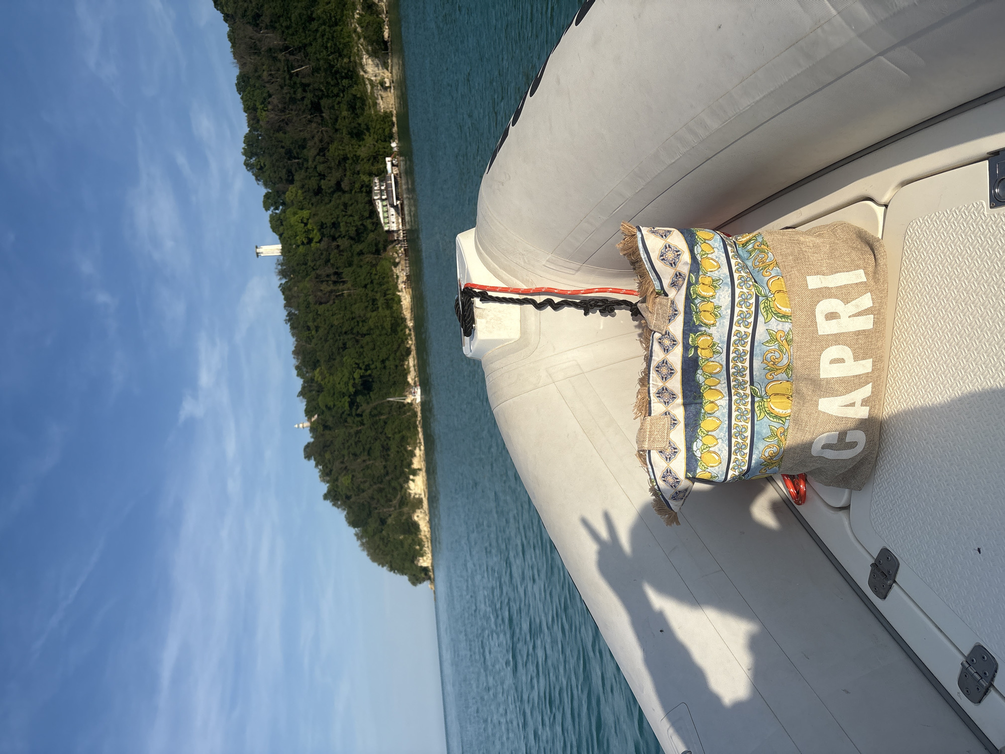The image presents a first-person perspective from the bow of a white inflatable boat moving across calm, clear turquoise water. A light brown, coarse-fabric bag, printed with the word "CAPRI" in white letters and adorned with a blue, yellow, and white lemon and tile pattern, rests on the boat's textured deck in the foreground. A red and black rope is coiled and secured to a white fixture at the boat's bow. A shadow, depicting a hand making a peace sign, is cast on the interior of the boat's port side.
In the midground, a dense, green forested landmass, characterized by a light-colored rocky shoreline, extends into the water. Further into the background, two distinct white, cylindrical tower structures, resembling lighthouses, are visible above the tree line on the elevated terrain. A multi-story white building with a dark roof is situated close to the waterline on the far right of the landmass. A small white sailboat is anchored offshore to the left of the main landmass. The sky above is light blue with scattered wispy clouds. The scene is illuminated by natural daylight, casting shadows to the right. The location is Unknown, Unknown.
