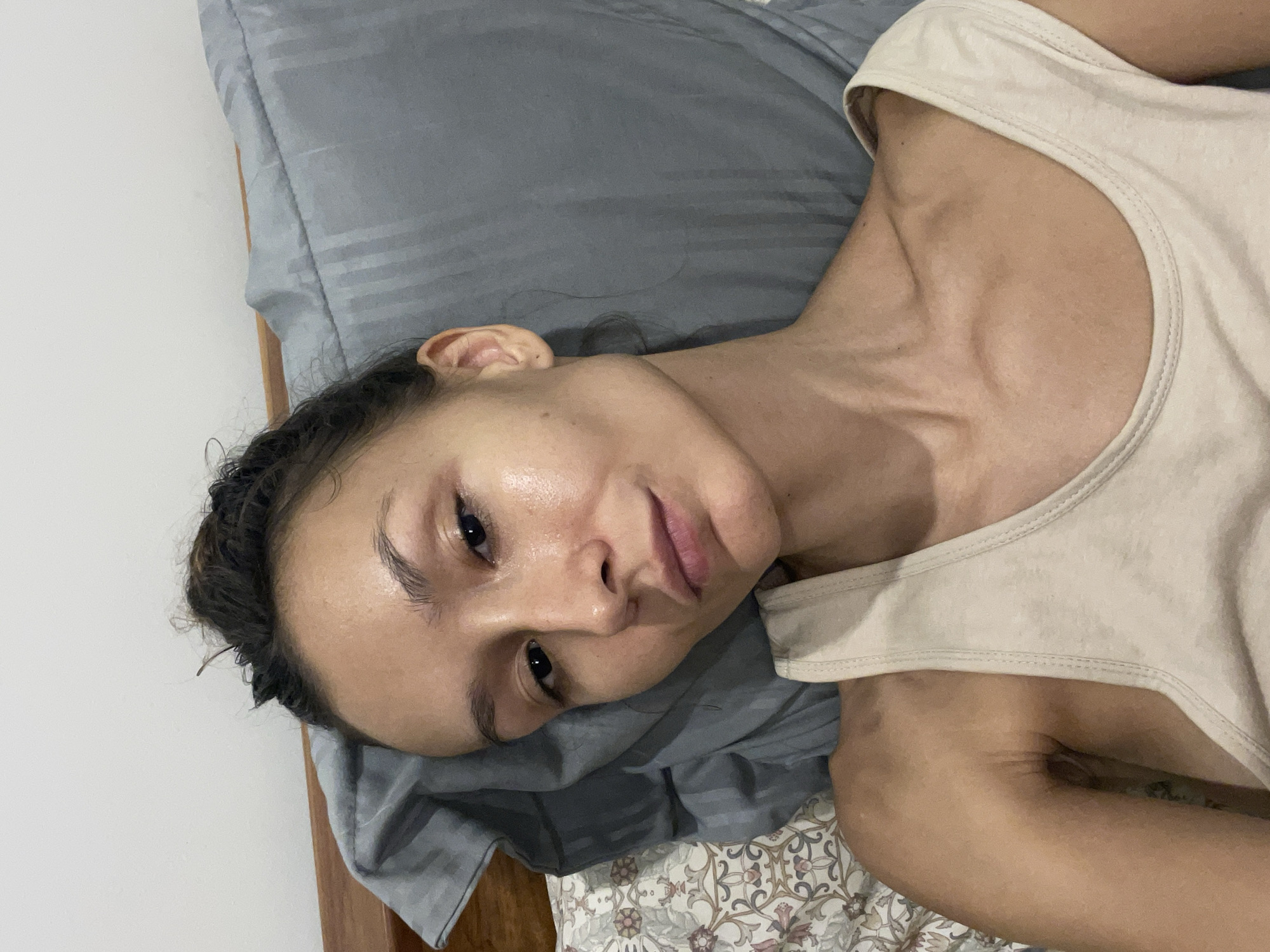 A young adult female is depicted in a medium close-up, lying on her side in bed, looking towards the camera. Her head rests on a gray pillow, with her body oriented to the right of the frame (if the image were rotated to portrait). She has dark, curly hair, appearing to be pulled back or in a loose braid. Her skin has a natural, slightly dewy sheen. Her eyes are open and dark, with a calm, neutral, or slightly pensive expression. Her lips are a natural pink hue.
She is wearing a simple, light-colored (beige or off-white) sleeveless tank top. Prominent veins are visible on her neck and shoulder area, indicating a lean physique. The bed features a plain gray pillowcase and a decorative bedsheet with a floral or intricate pattern in muted tones, visible beneath the pillow. A plain white wall serves as the background on the left side of the frame. The lighting is soft and even, suggesting an indoor setting, possibly a bedroom. The overall mood is relaxed and quiet.
This scene is contextually located in Mũi Né, Vietnam, though no specific visual cues confirm this within the image itself. There is no visible text.