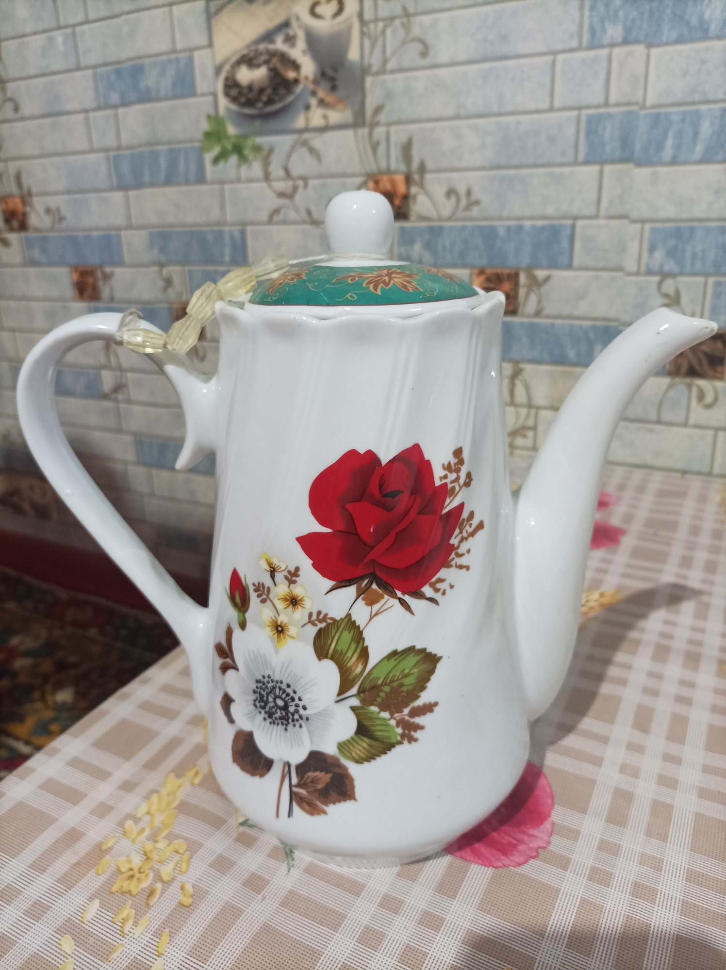 A white ceramic teapot with a floral design, featuring a red rose and white flowers, sits on a checkered tablecloth. The teapot has a white handle and spout, and a green lid adorned with a floral pattern. A light yellow beaded ribbon is tied around the neck of the teapot. The background features a tiled wall with blue and white patterns. The scene appears to be indoors, possibly in a home in Seýdi, Turkmenistan.