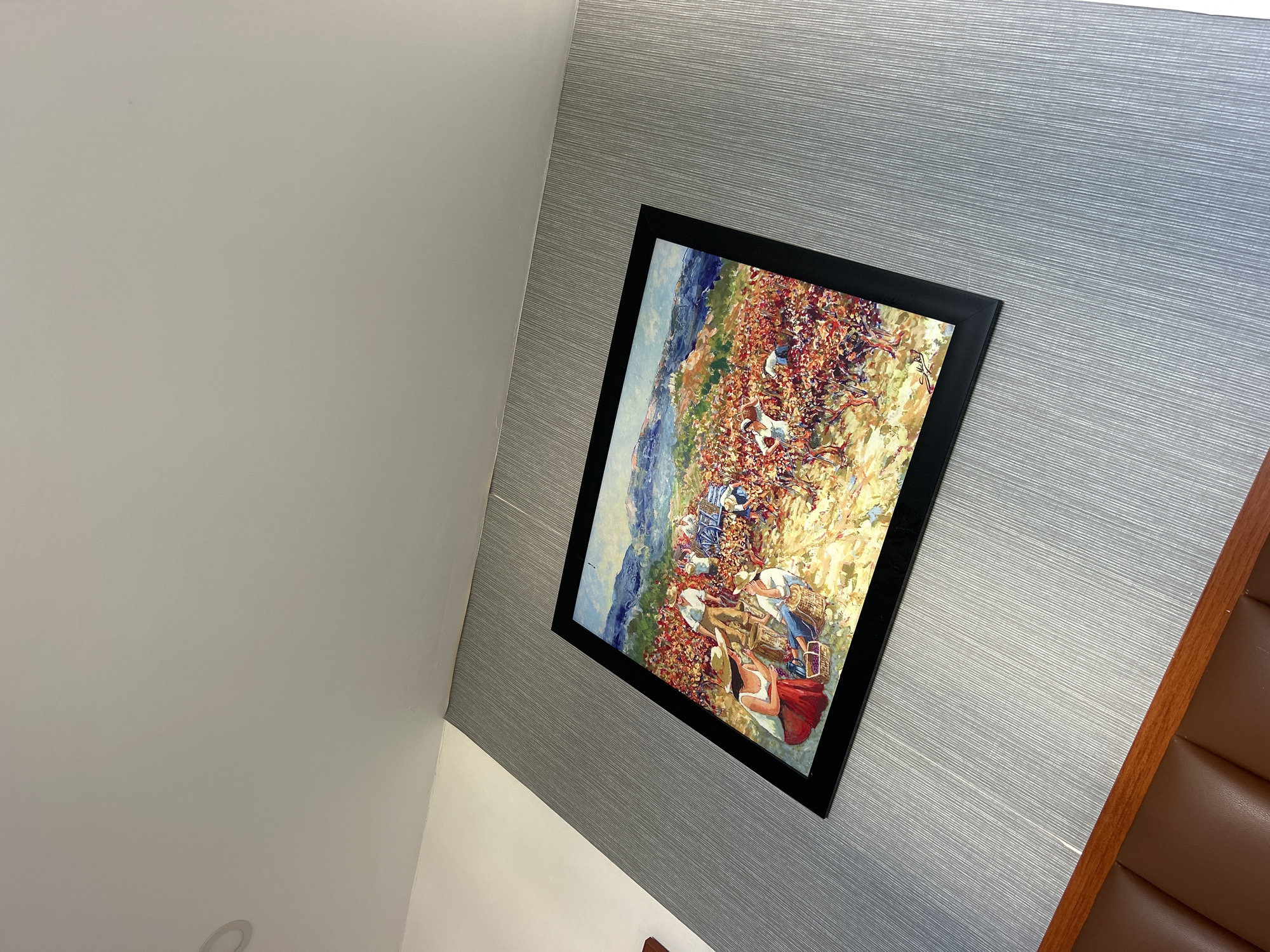 A framed painting hangs on a gray wall. The painting depicts a group of people working, possibly in a vineyard. The wall is gray with a horizontal texture. To the left of the wall is a white wall. To the right of the painting, a brown cushioned structure is visible. The image was taken indoors. Location is unknown.