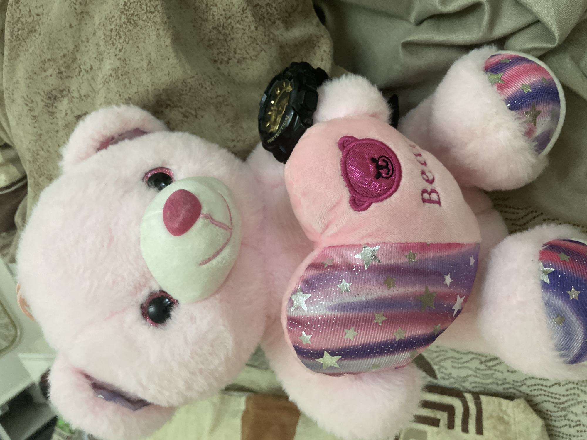 This image captures a pink teddy bear lying on a textured light brown or beige fabric, possibly bedding, in Ihtiman, Bulgaria. The teddy bear is the central subject, rendered in a soft, fluffy pink material.
Notable details of the teddy bear include its black, sparkling eyes with pink glittery eyelids, and a white snout with a bright pink nose and a stitched smile. A black digital watch, resembling a G-Shock, is fastened around one of the bear's arms; the word "PROTECTION" is clearly visible on its casing, and it features gold-colored internal components.
On the bear's chest, there's a shimmering pink patch embroidered with a cartoon bear's head logo and the word "Bear" written vertically in a darker pink or maroon shade. The bear's four paws feature decorative patches with a vibrant, swirled pattern of pink, purple, and blue hues, accented with numerous silver glittery stars. The overall scene suggests a cozy, indoor setting.