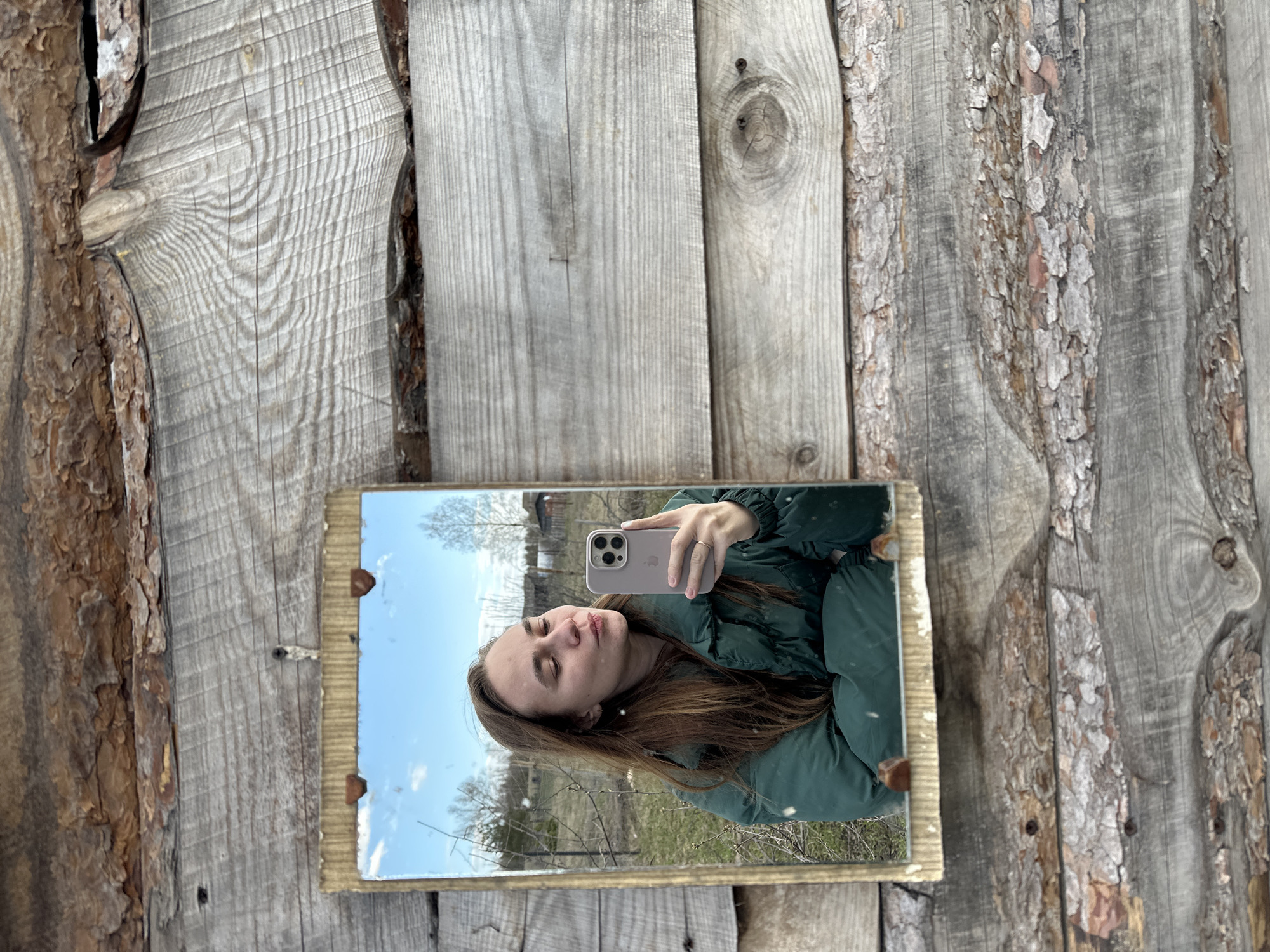 A woman is depicted taking a selfie in a mirror, which is mounted on a rustic wooden surface outdoors. The woman, who has long brown hair, is wearing a dark green puffer jacket. She holds a light-colored iPhone, displaying an Apple logo on its case, to capture her reflection in the rectangular mirror. Her eyes are partially or fully closed, and her lips are slightly pursed in a posed expression.
The mirror reflects a clear blue sky with a few scattered clouds and a sparse landscape featuring bare trees, indicative of early spring or late autumn. A distant rural scene, possibly including a building, is also visible in the reflection. The mirror has a simple, distressed-looking frame, possibly made of wood or cardboard.
The wooden surface behind the mirror appears to be a weathered fence or wall, constructed from rough-hewn planks. Many of these planks exhibit natural bark edges, enhancing the rustic feel. The wood grain is prominent, and some knots and screws are visible in the planks. The setting suggests an outdoor, rural environment, consistent with Kharnavy, Russia, under bright daylight conditions.
