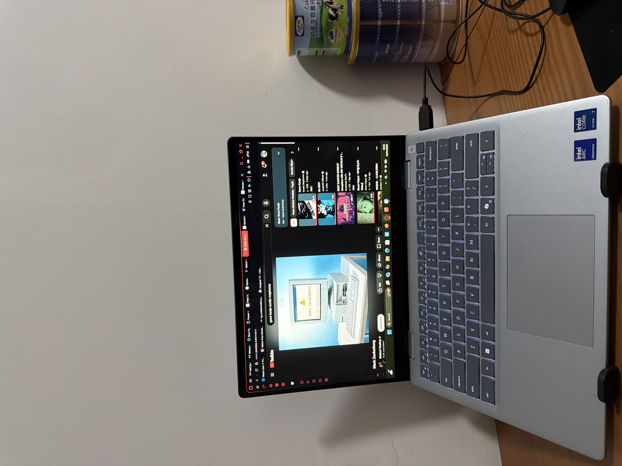 A silver laptop sits on a wooden surface next to a blue can. The laptop screen displays a YouTube page with various music videos and a video titled "MARK ZUCKERBERG". The laptop has Intel Arc graphics and Intel Core Ultra 7 stickers on the keyboard side. The can appears to be a food product, possibly a milk powder. A black cable is plugged into the side of the laptop. The background shows a plain white wall. It is not possible to determine if the image was taken in Jiangning District, China, with the available information.